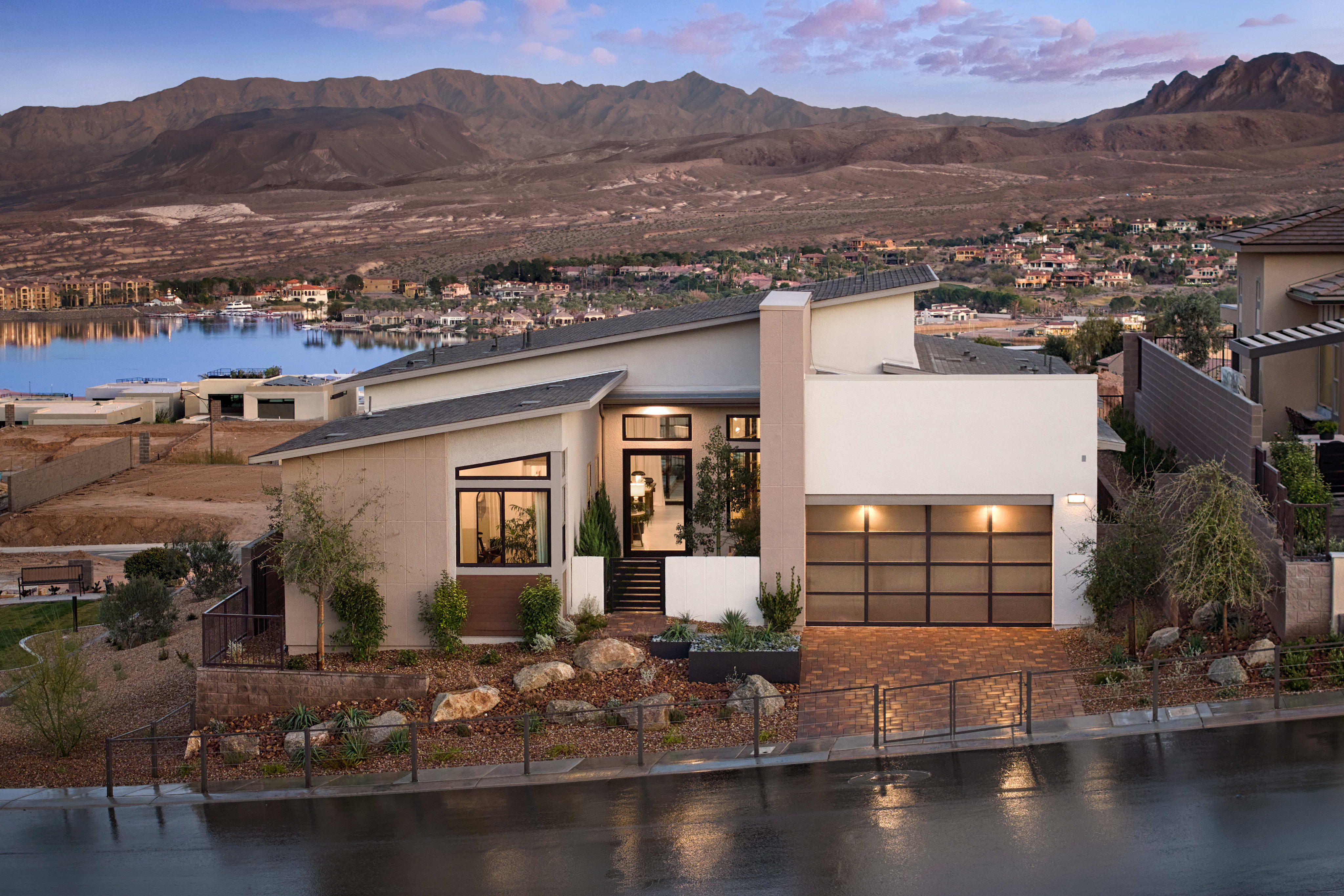 A modern, multi-level house nestled on a hillside overlooking a scenic lake and mountainous landscape in the background.
