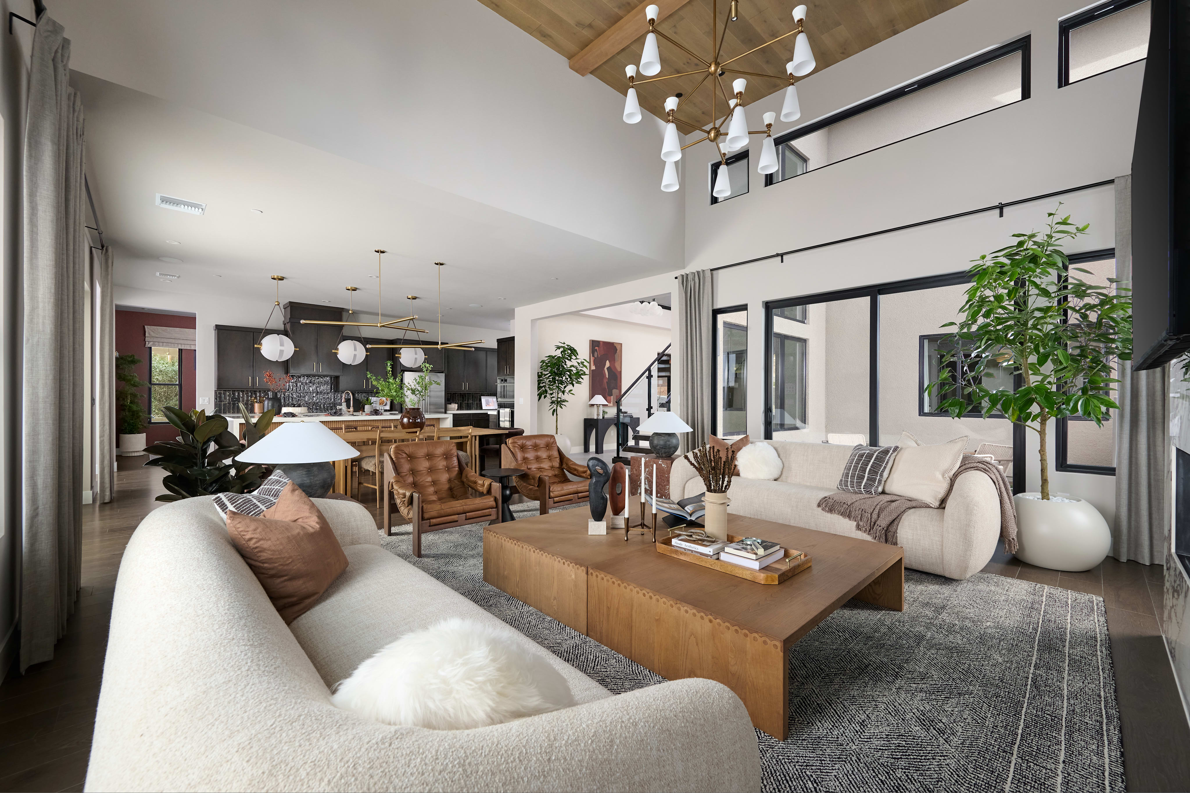 A spacious and modern living room with a cozy seating area, a wooden coffee table, and a kitchen visible in the background, all illuminated by a stylish chandelier.