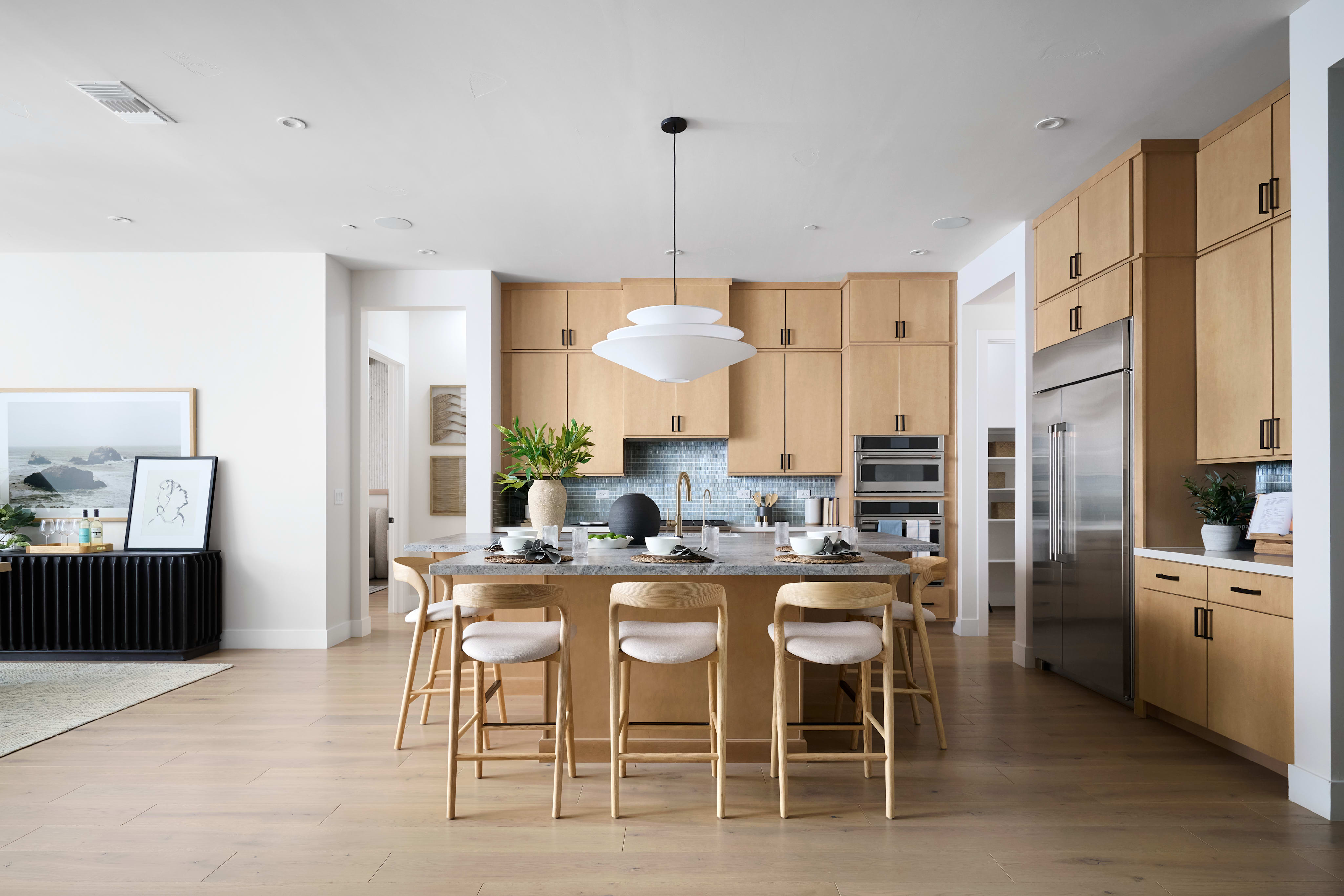 A modern, open-concept kitchen with light wood cabinetry, a large island with bar stools, and a dining area visible in the background.