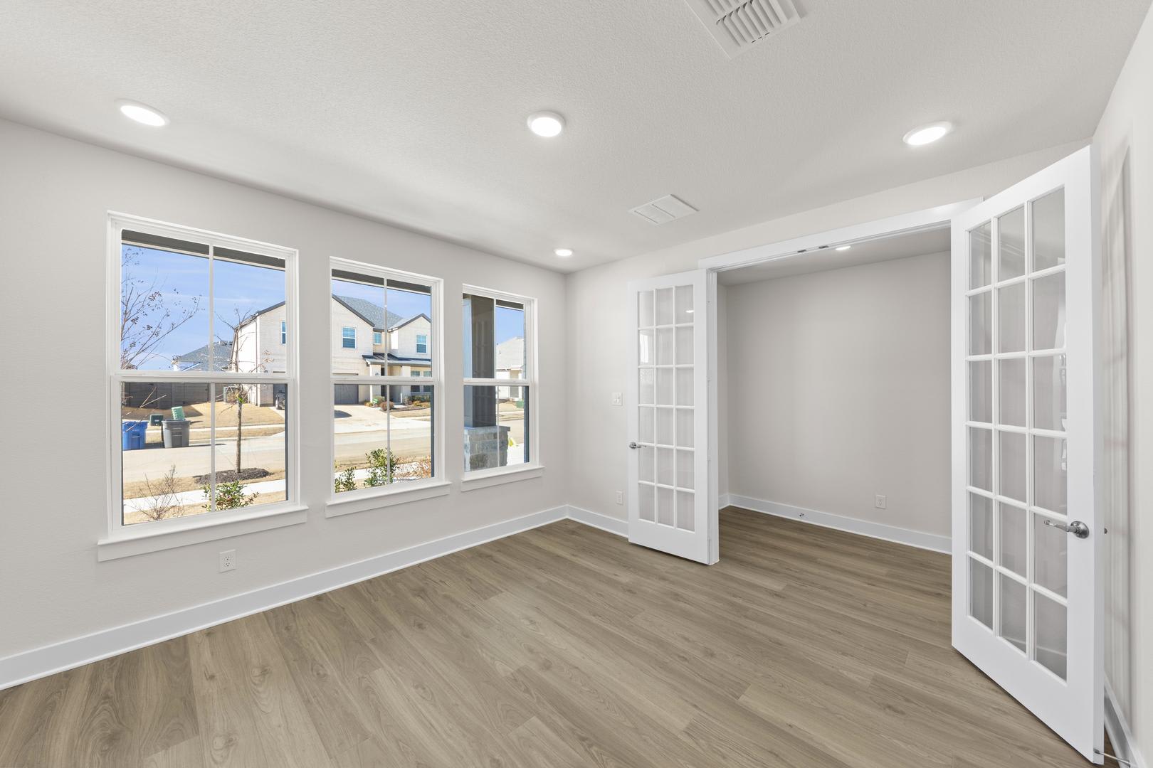 A bright and spacious room with large windows, white walls, and a wooden floor. The room appears to be a living or dining area, with a French door leading to the exterior.