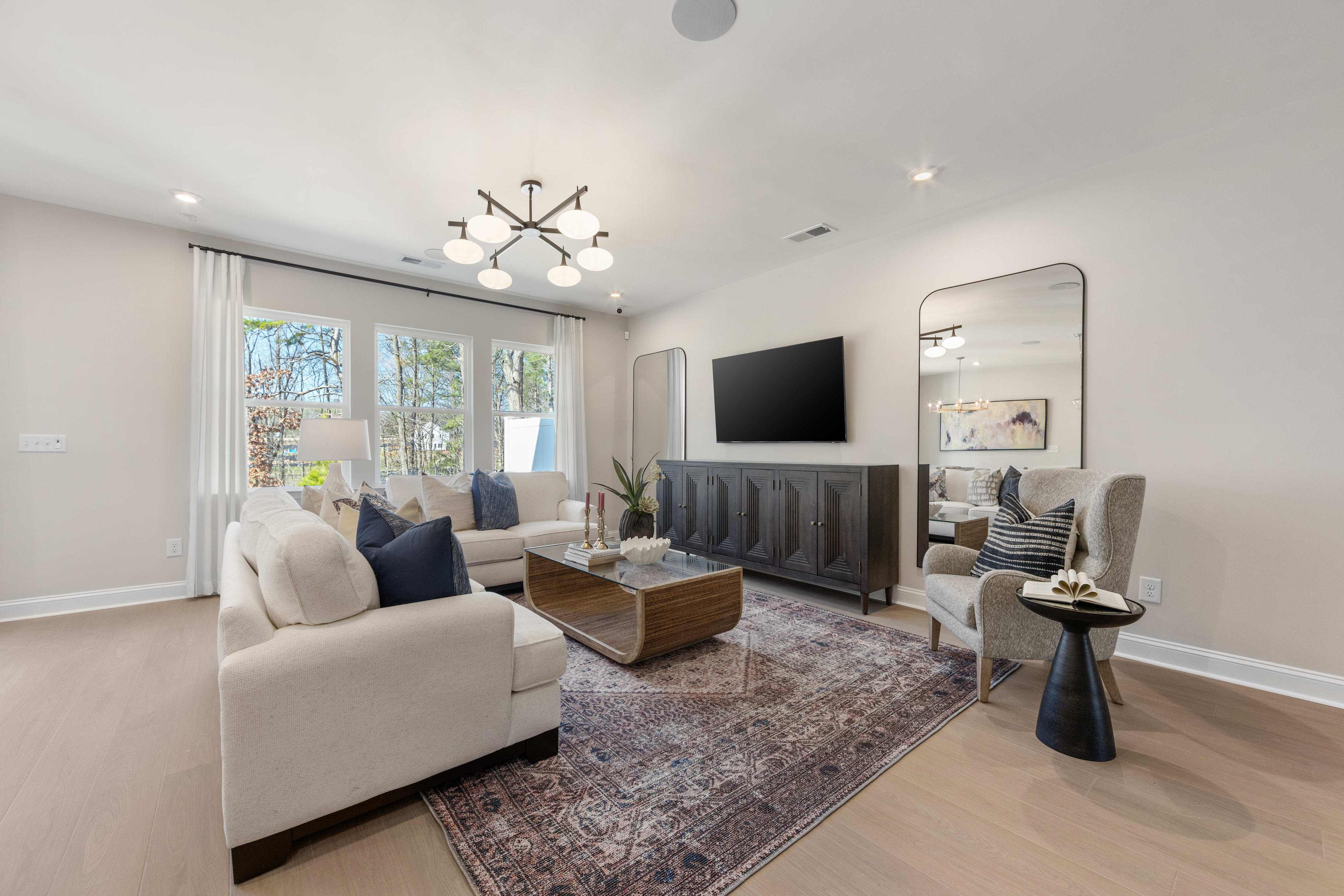A modern and stylish living room with a large TV, comfortable seating, and a decorative chandelier overhead, set against a backdrop of large windows and a neutral color scheme.