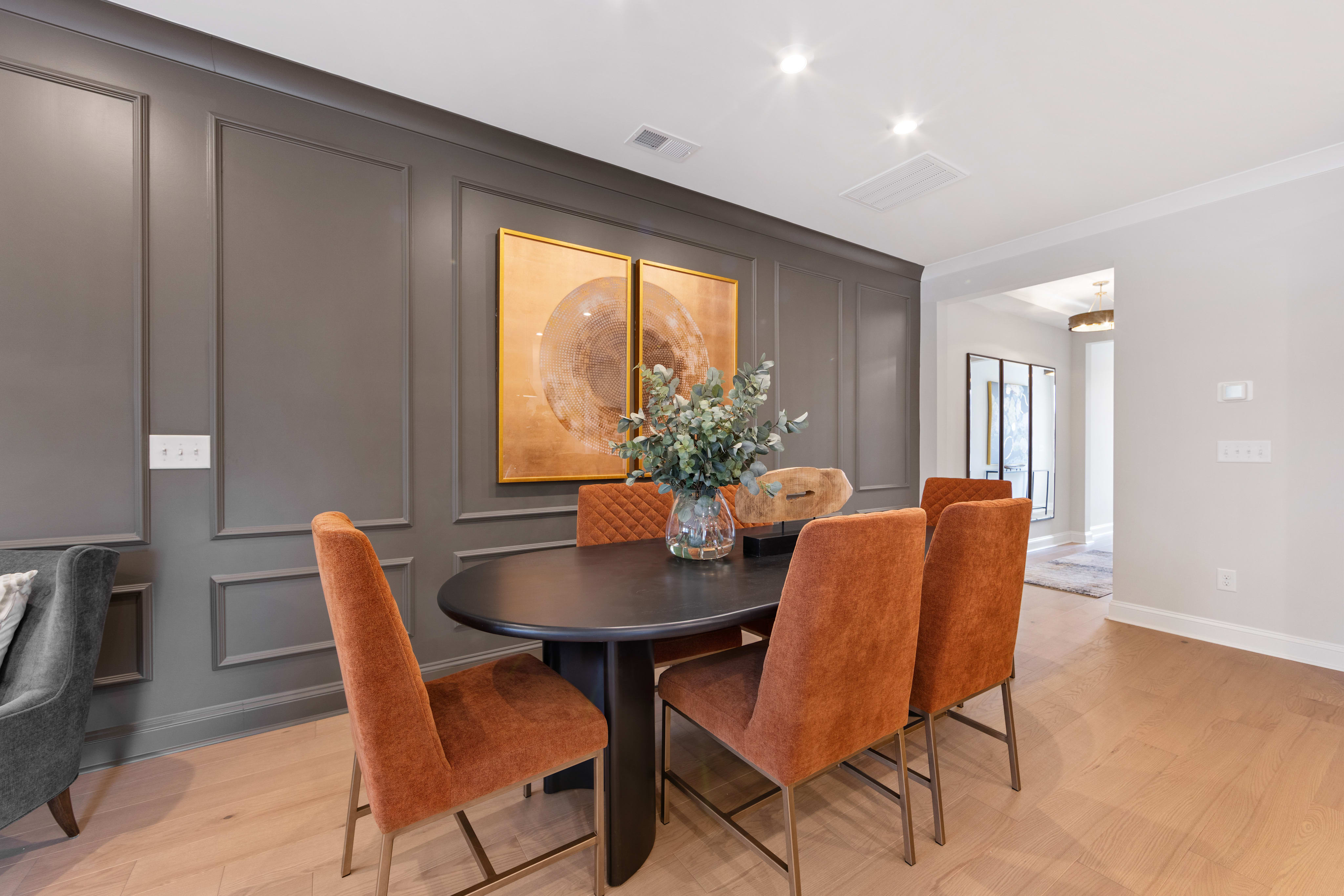 A modern dining room with a dark gray accent wall, a round wooden table, and orange upholstered chairs, complemented by a floral artwork and a vase of flowers.