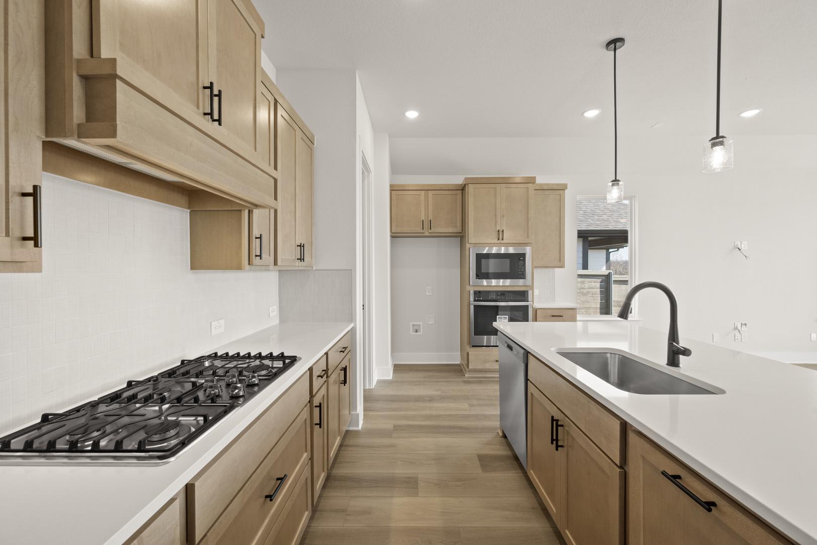 A modern, well-lit kitchen with light-colored cabinets, a gas stove, and a sink with a black faucet, set against a backdrop of white walls and pendant lighting.