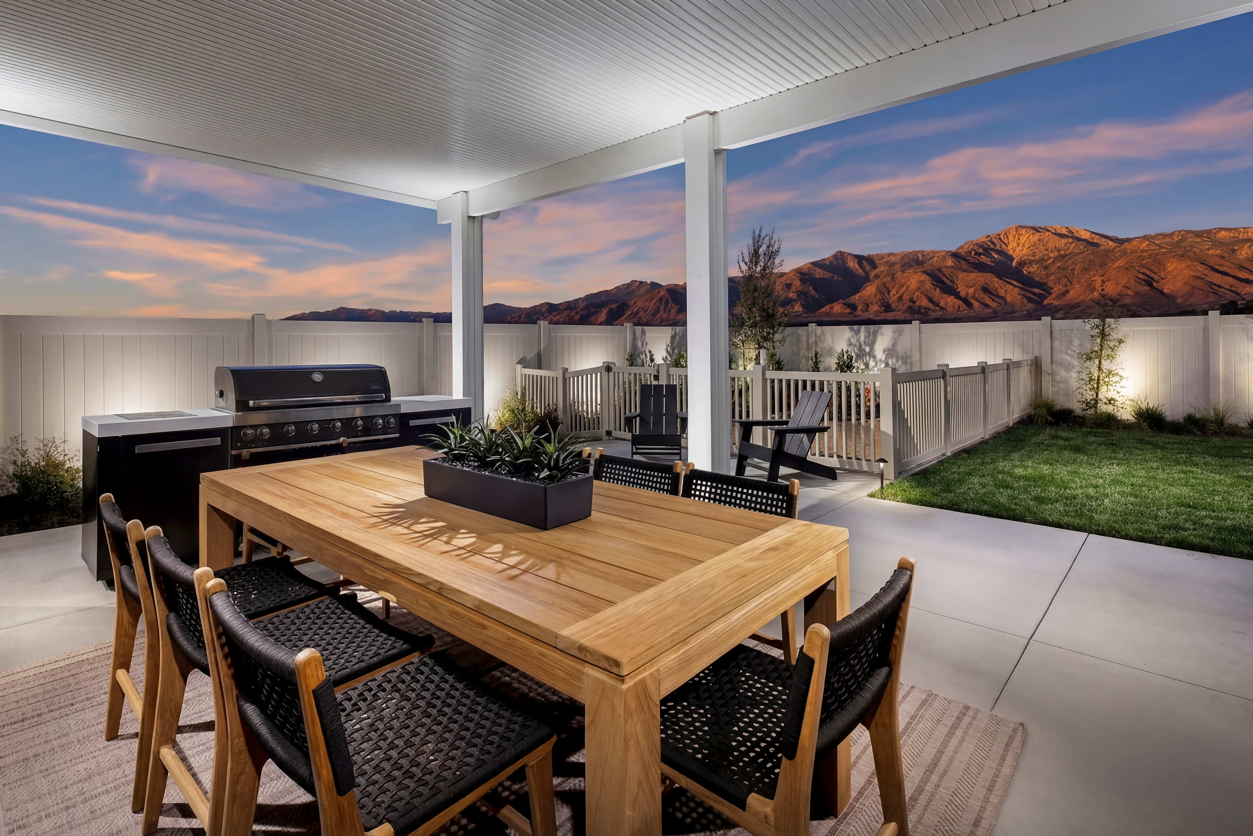 A modern outdoor dining area with a wooden table and chairs, surrounded by a lush green lawn and framed by a stunning mountain landscape in the background.