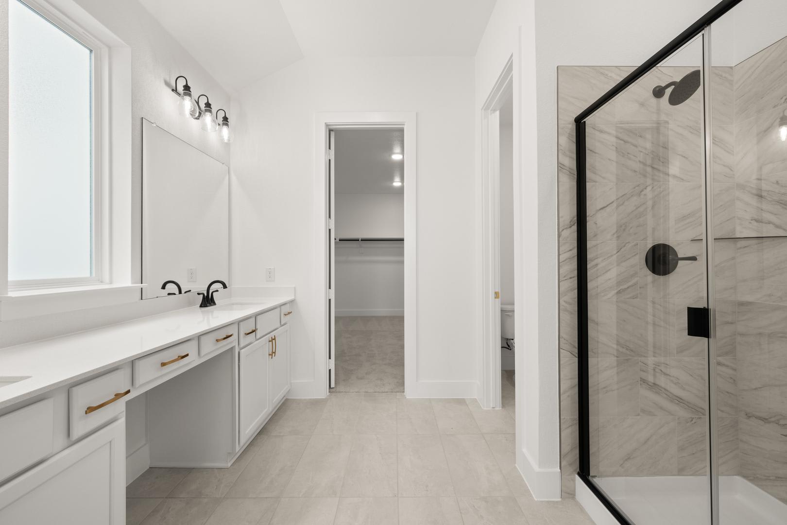 A modern, minimalist bathroom with a long vanity, a glass-enclosed shower, and a doorway leading to an adjacent room.