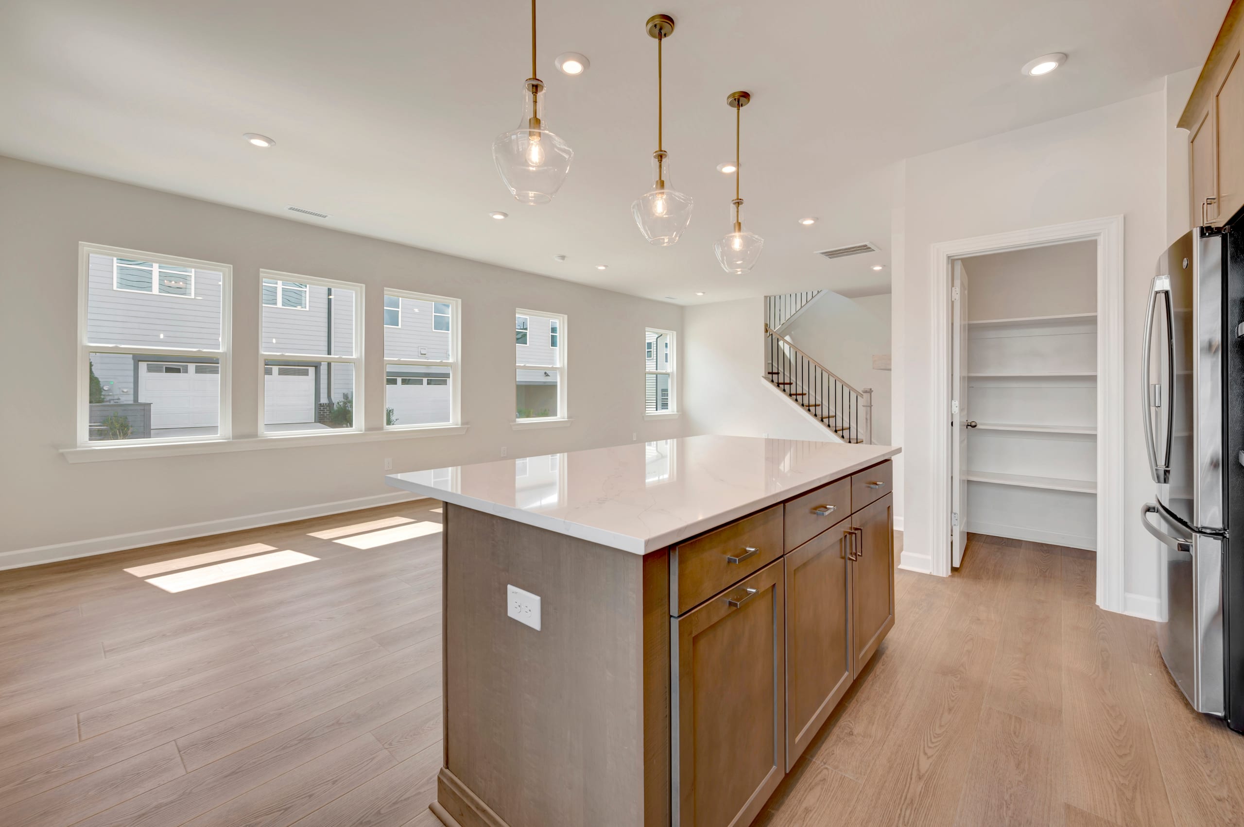 A modern, open-concept kitchen with a large island, wooden cabinets, and pendant lighting fixtures, set against a bright and airy background with large windows.