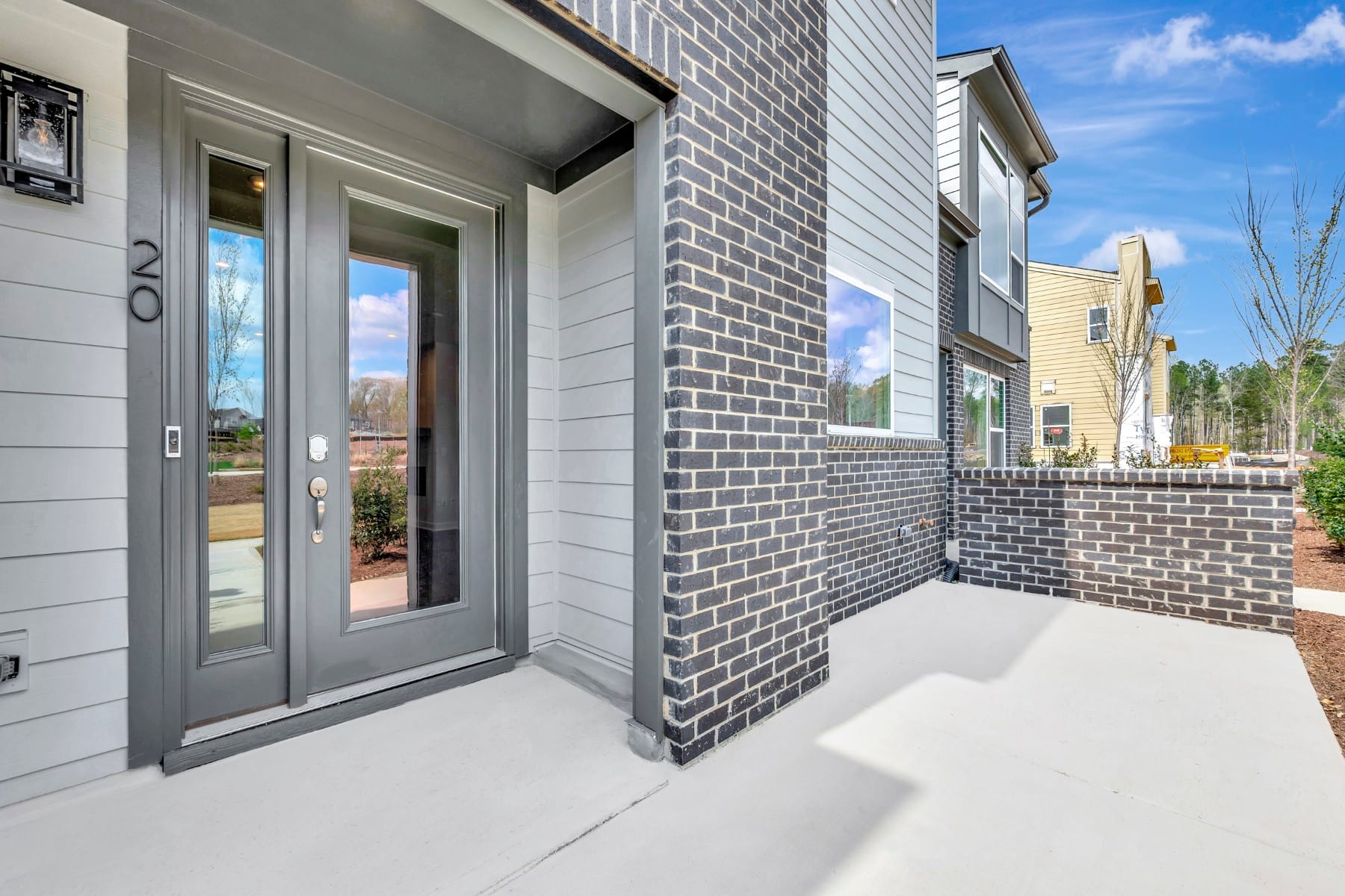 A modern, gray-colored townhouse with a brick exterior, a glass front door, and a paved walkway leading to the entrance, set against a backdrop of other similar buildings and a blue sky with clouds.