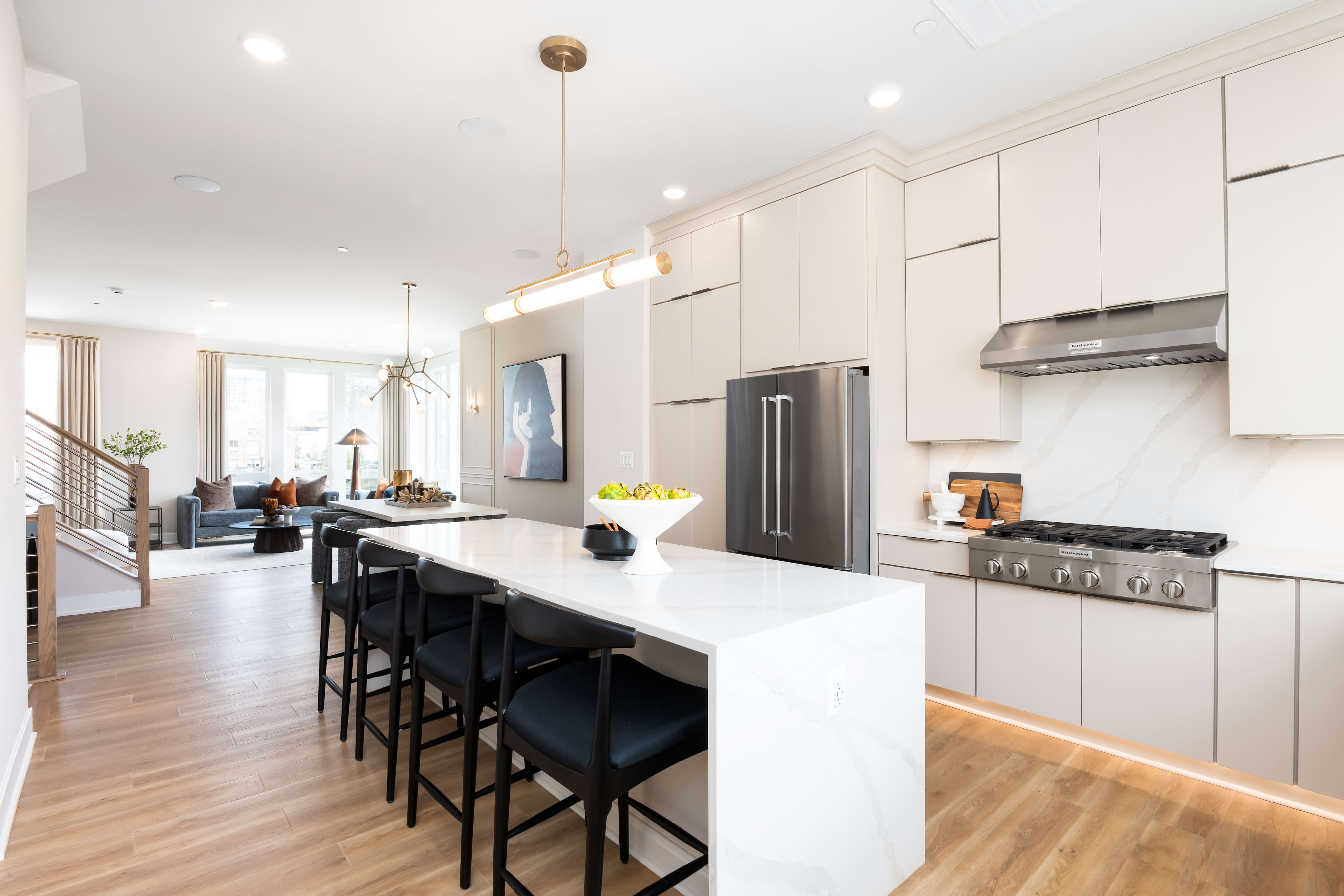 A modern, open-concept kitchen with white cabinets, stainless steel appliances, a large island with bar stools, and a hardwood floor leading into a living area.