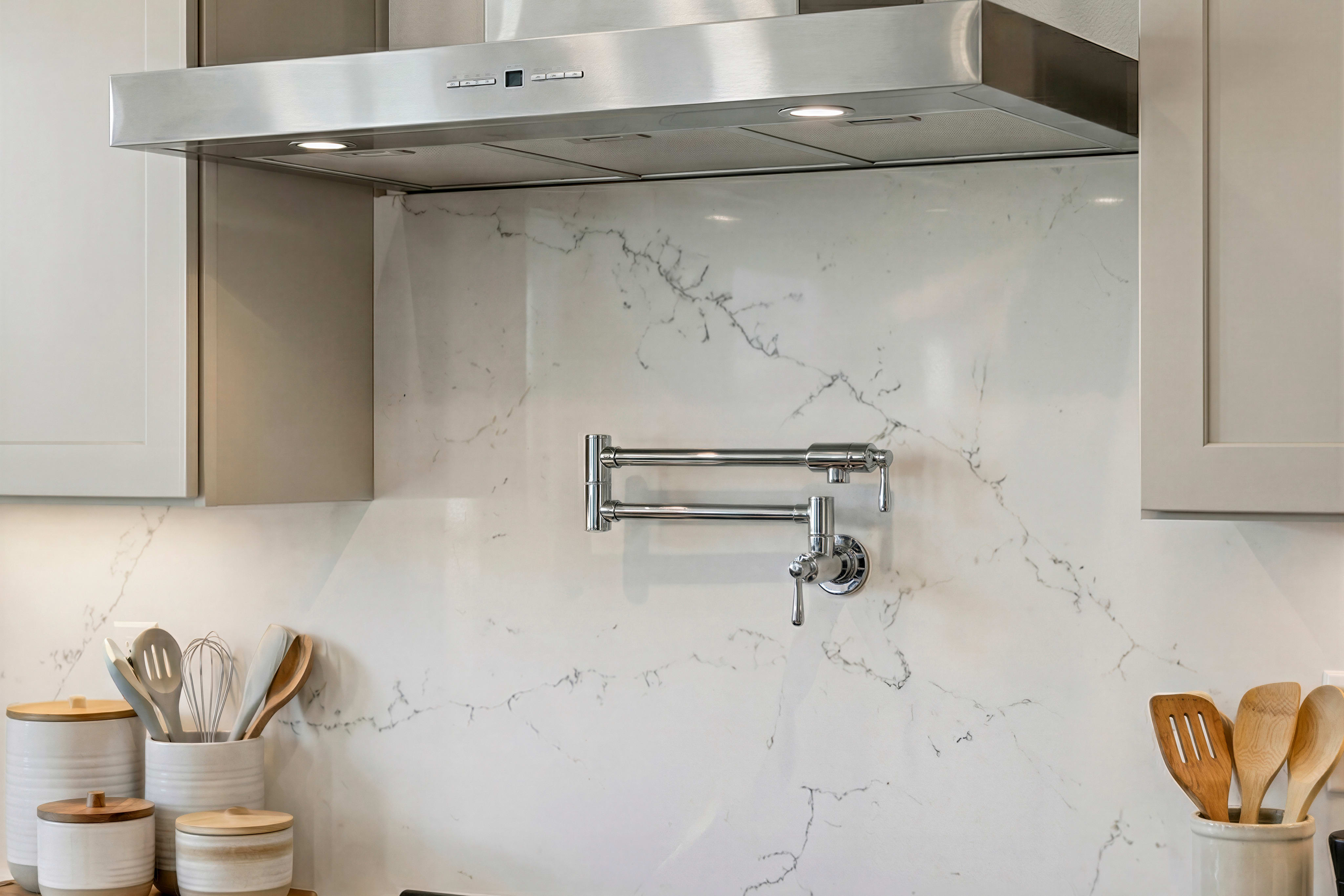 A modern, minimalist kitchen with a stainless steel range hood, a marble backsplash, and various cooking utensils neatly arranged on the countertop.