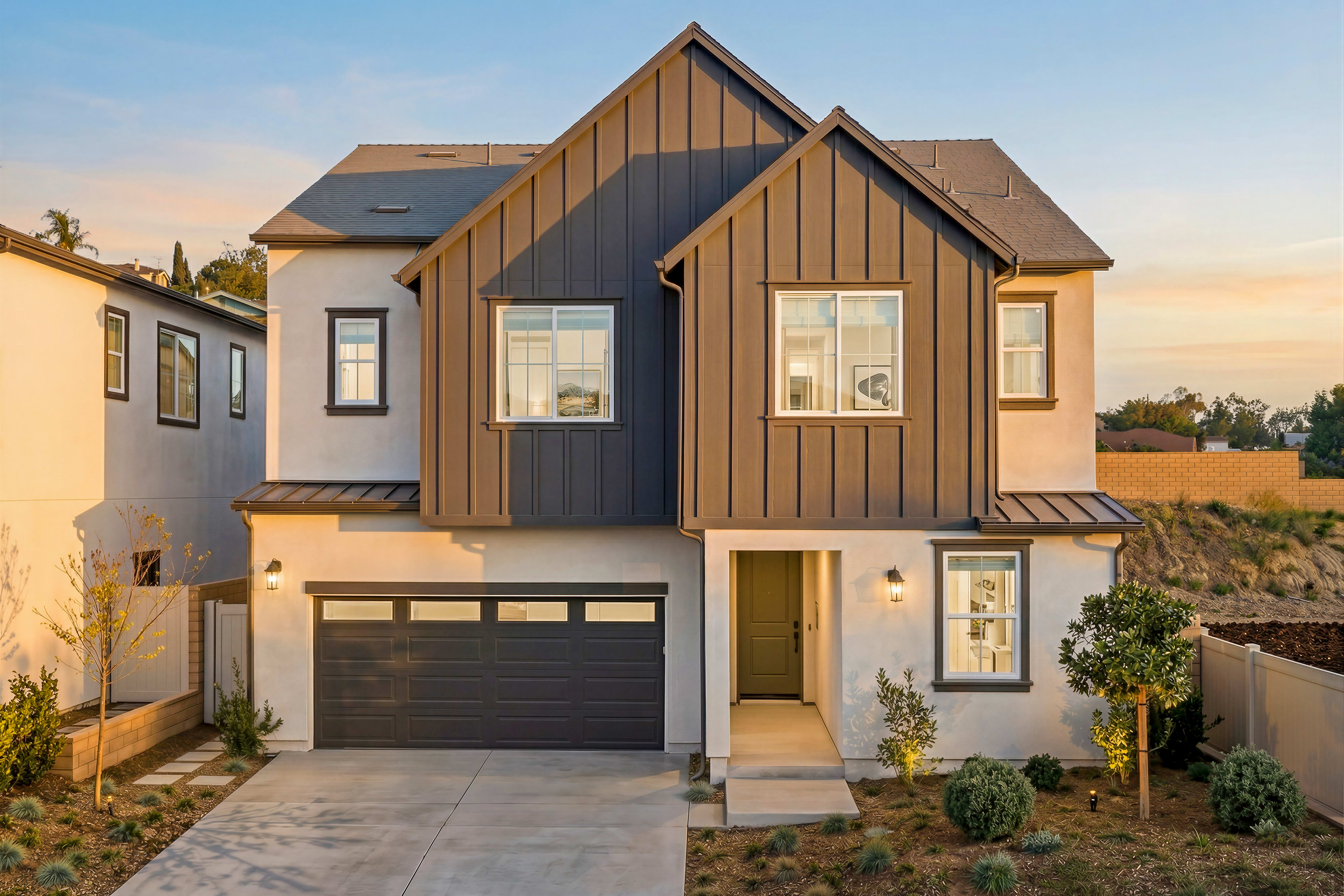 A modern two-story house with a mix of wood and gray siding, a garage door, and a paved driveway in the foreground, set against a backdrop of a clear sky and lush greenery.