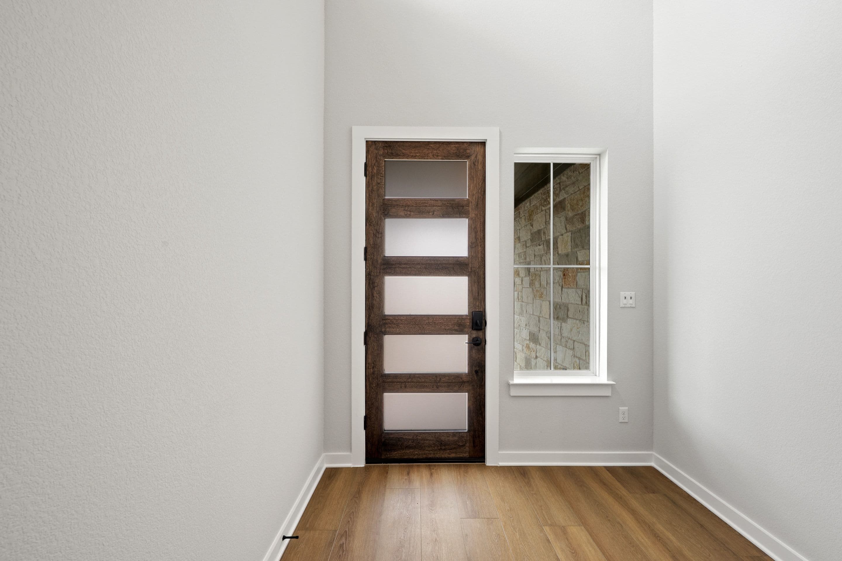 A wooden door with glass panels stands in a white-walled room, with a window visible in the background.