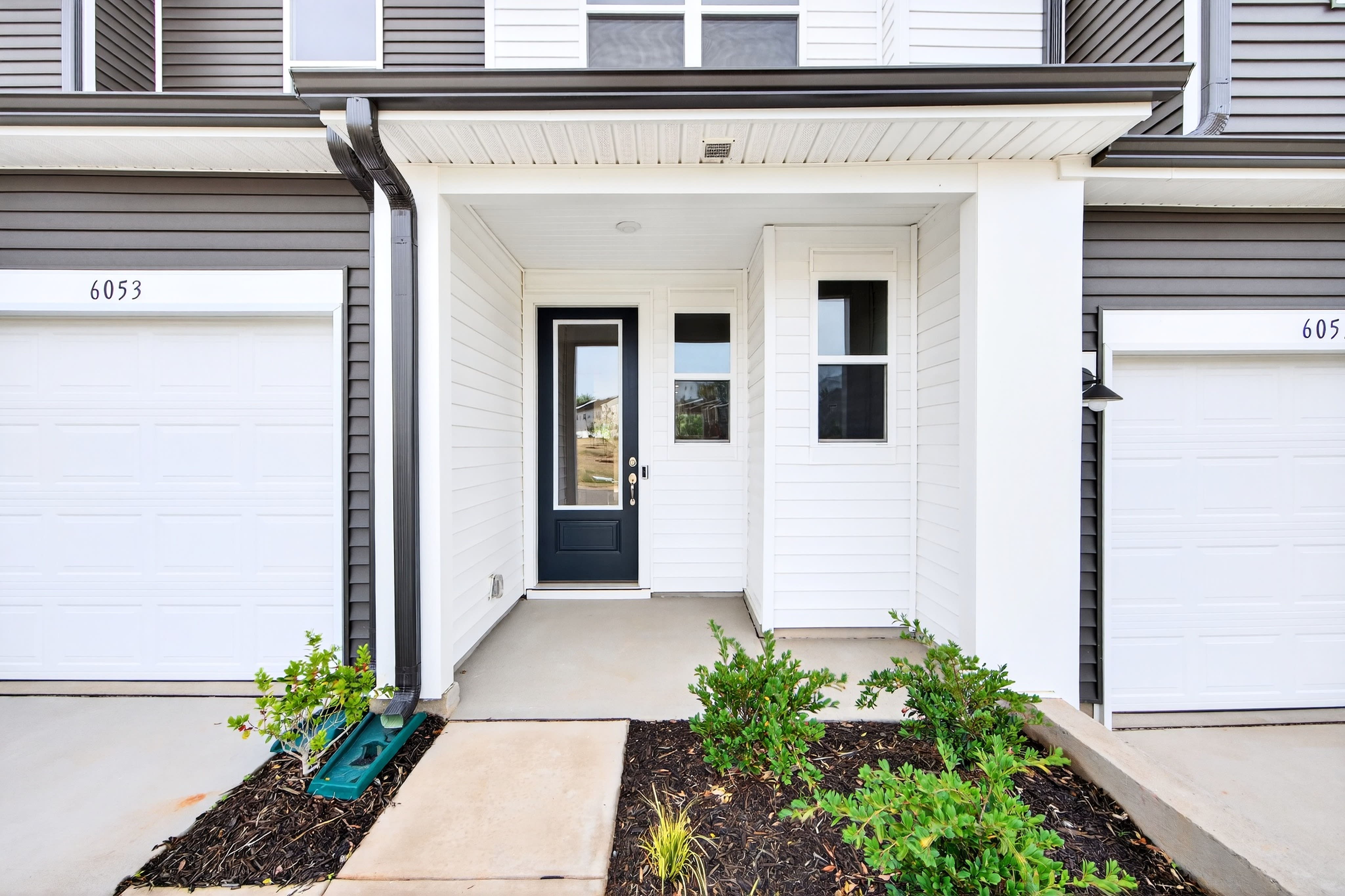 A modern, two-story residential building with a white exterior, a covered entryway, and a small garden bed in the foreground.