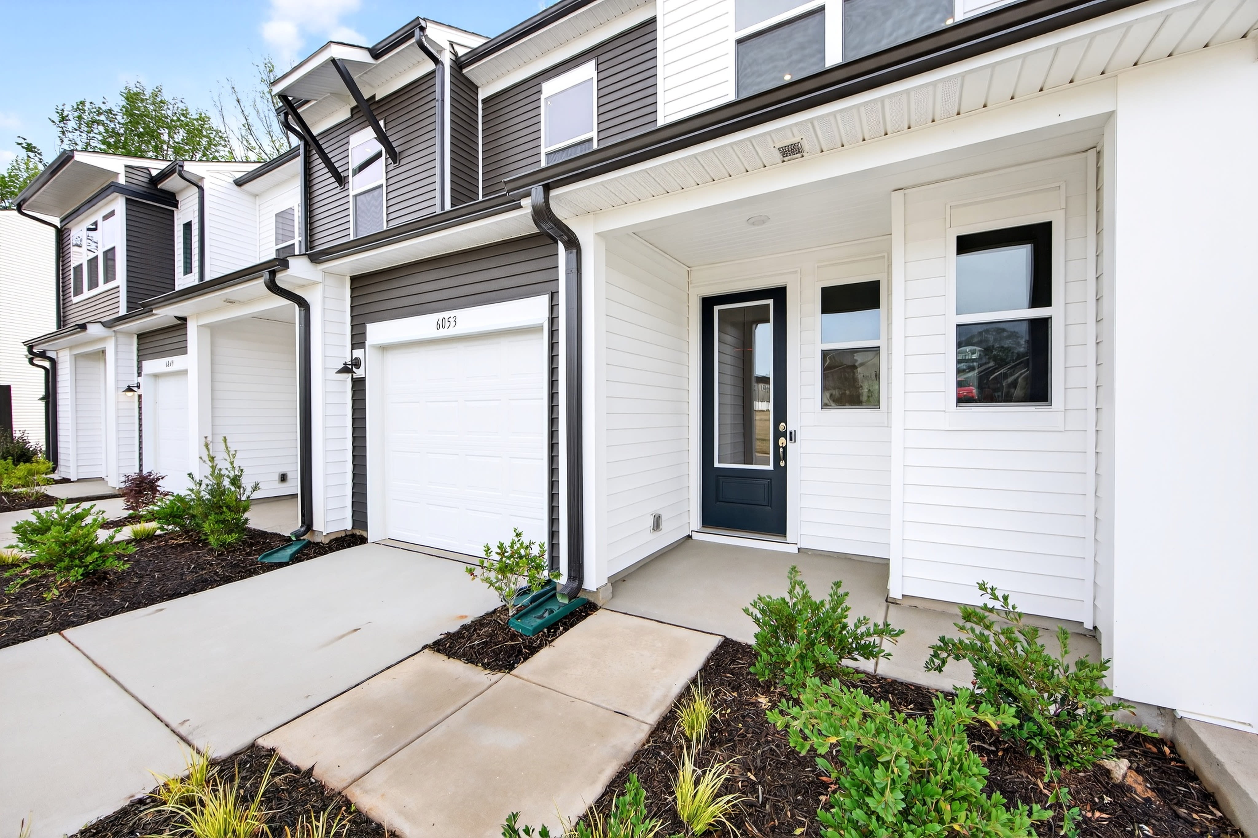 A modern townhouse with a well-landscaped front yard, featuring a concrete walkway leading to the white front door and surrounded by lush greenery.