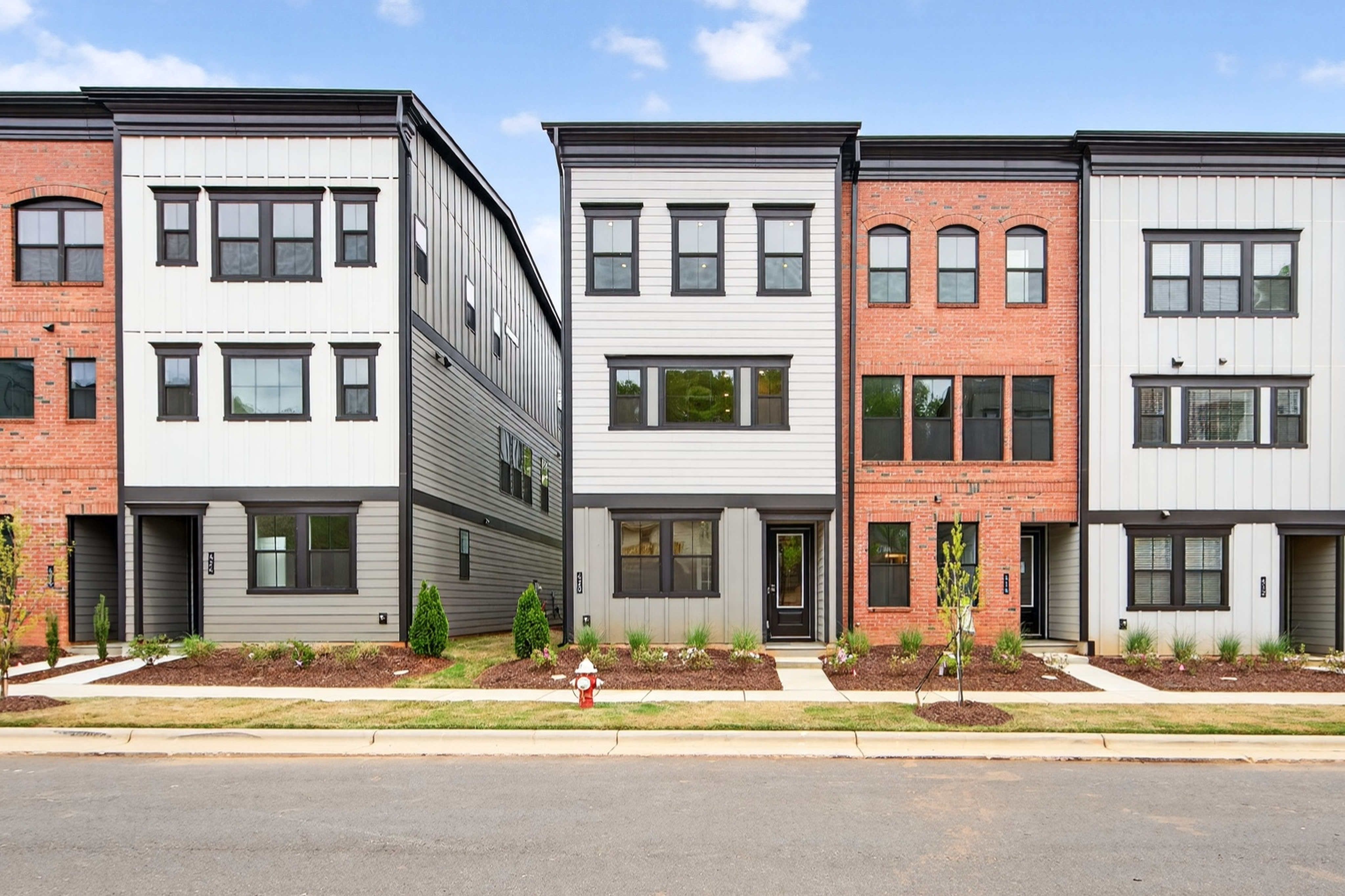 A row of modern, multi-story townhouses with a mix of brick and siding exteriors, surrounded by landscaped yards and a paved street in the foreground.