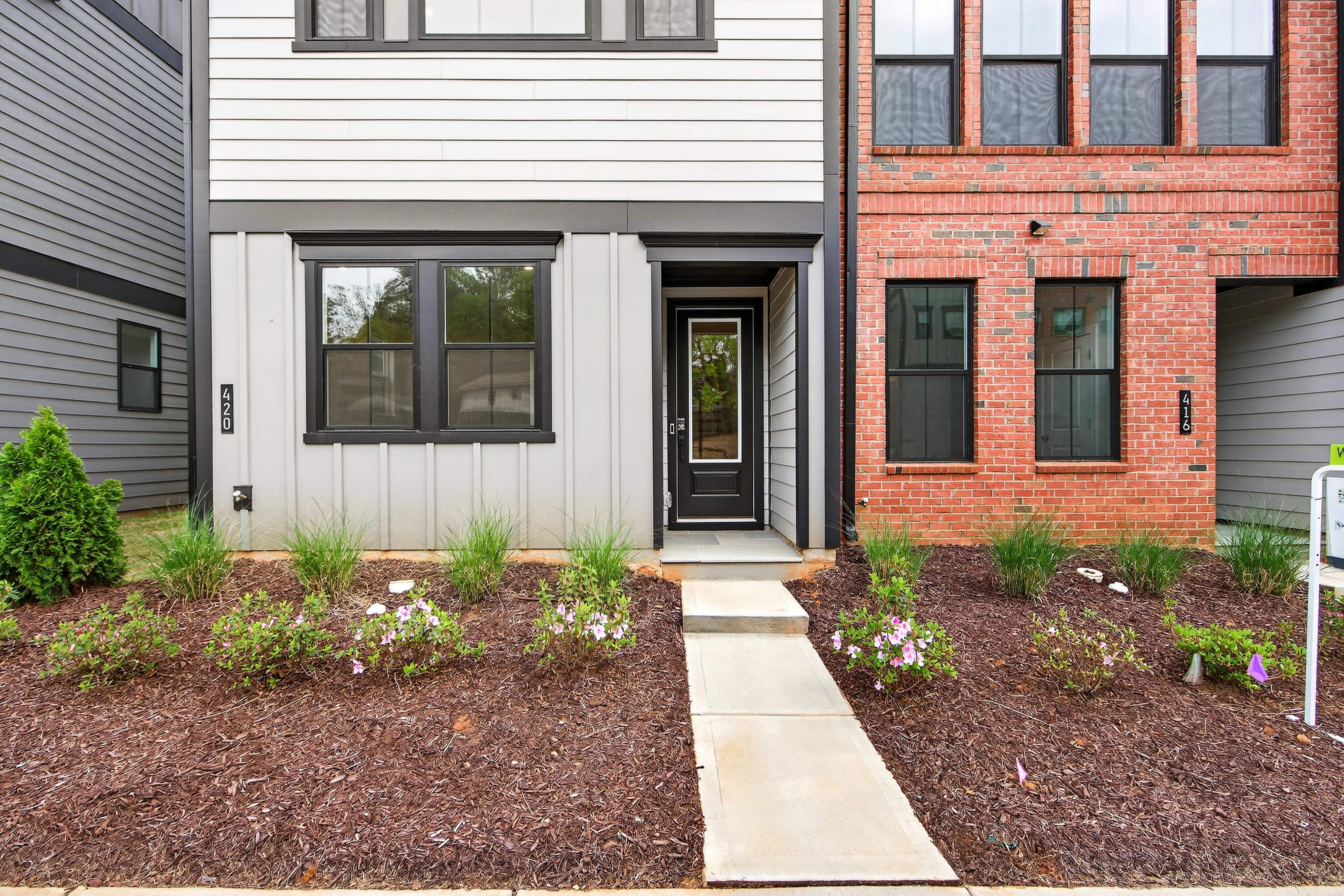 The image shows a modern townhouse with a brick exterior and a landscaped front yard featuring a concrete walkway leading to the entrance.