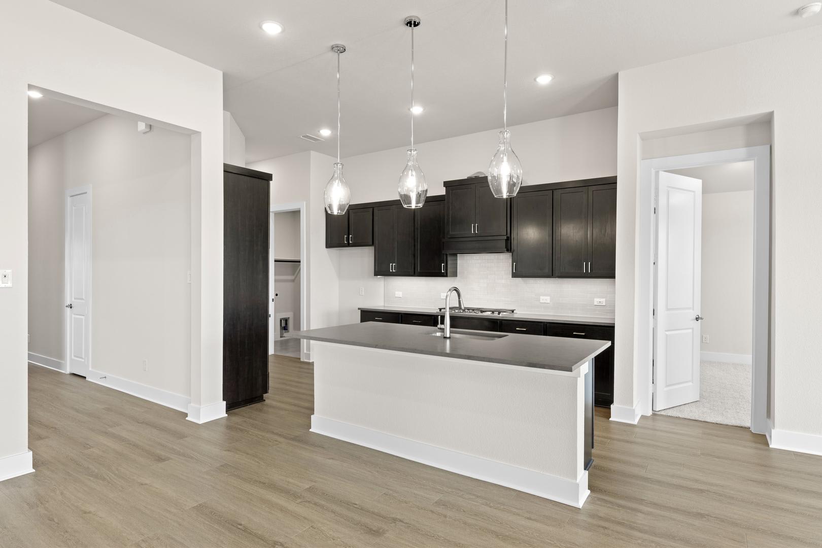 A modern, open-concept kitchen with dark cabinets, a white island, and pendant lighting fixtures, set against a light-colored hardwood floor.