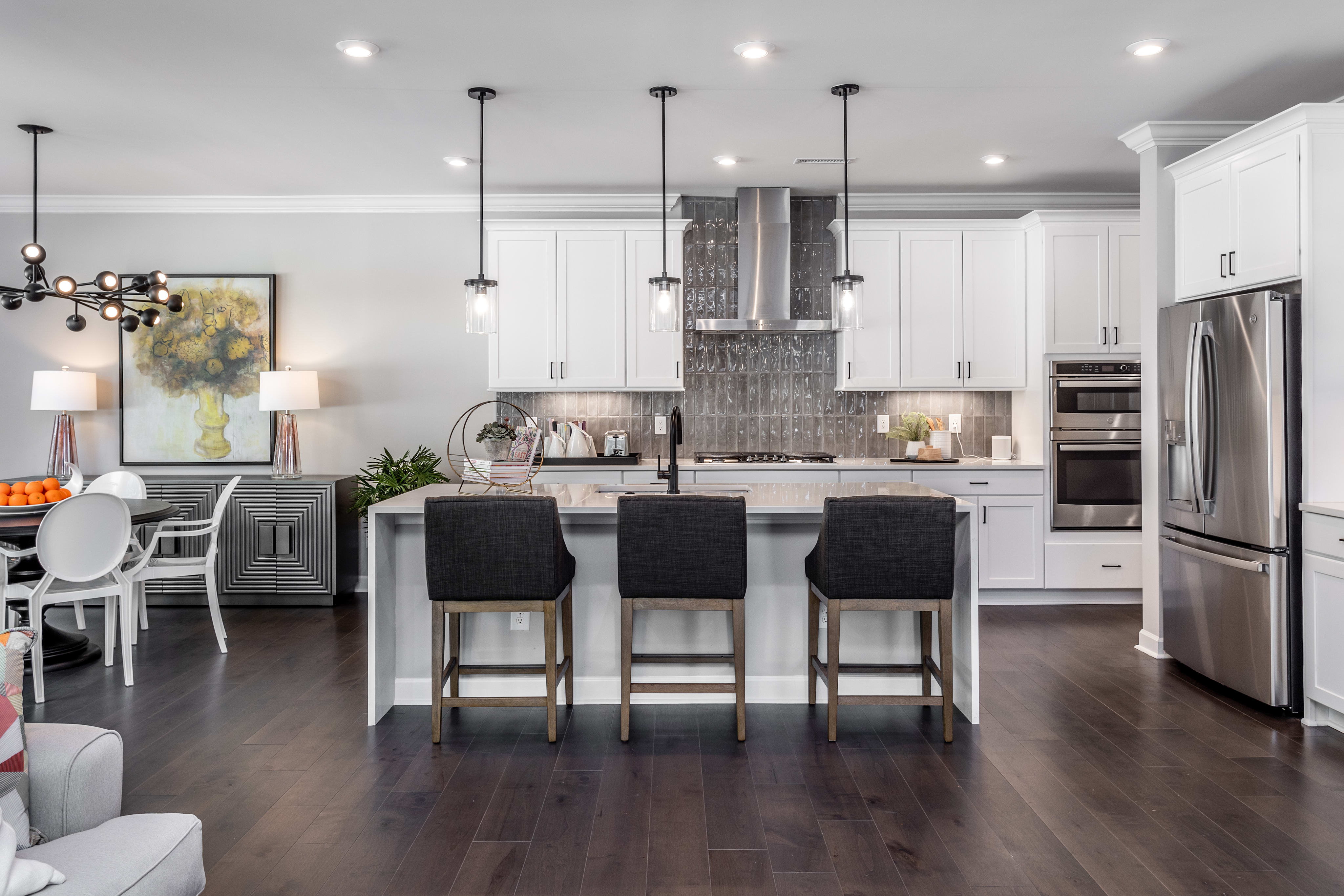 A modern, open-concept kitchen with white cabinets, stainless steel appliances, a large island with black bar stools, and a mix of pendant and recessed lighting creating a warm and inviting atmosphere.