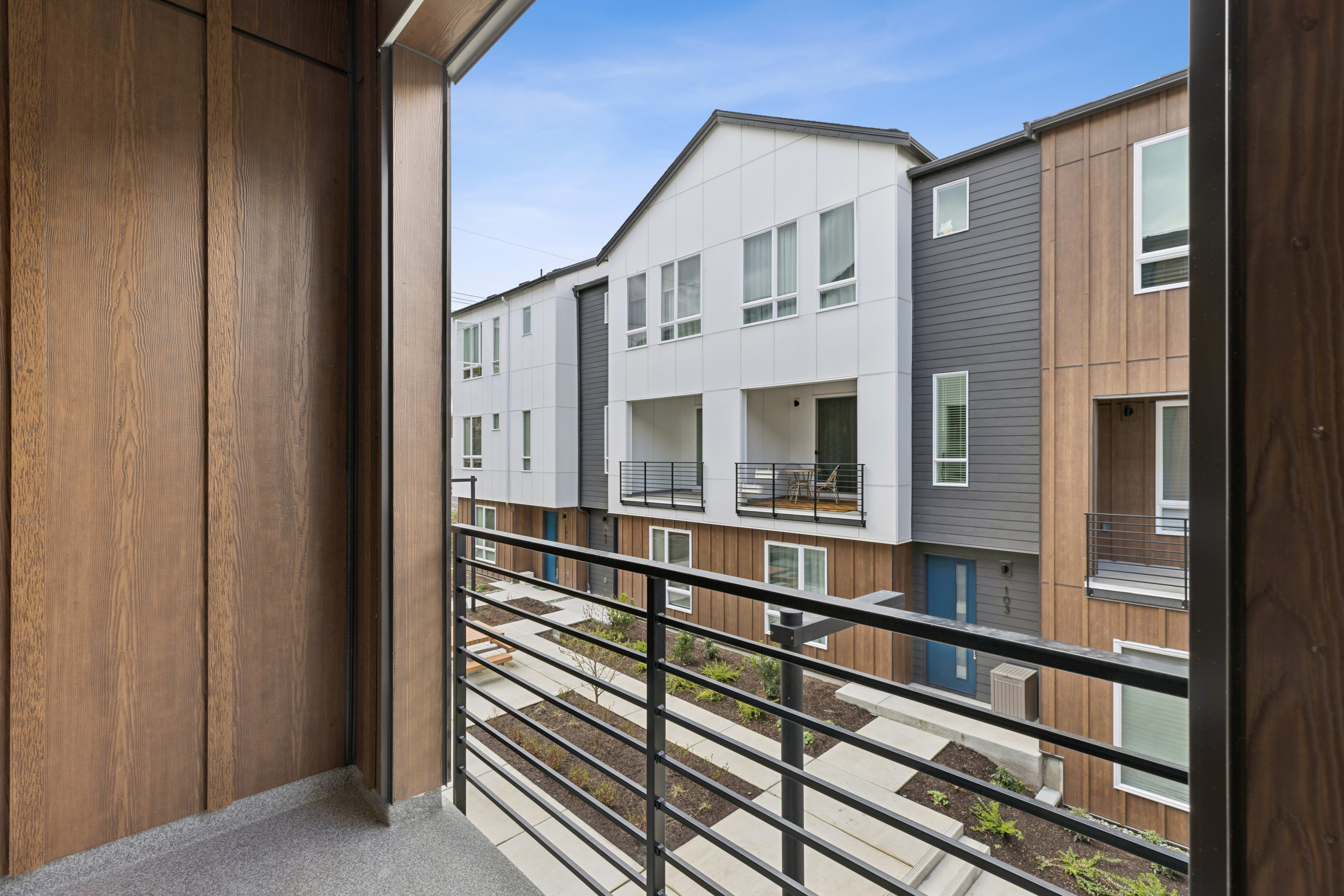 A wooden balcony with a metal railing overlooks a row of modern, multi-story residential buildings with a mix of siding materials and window styles.