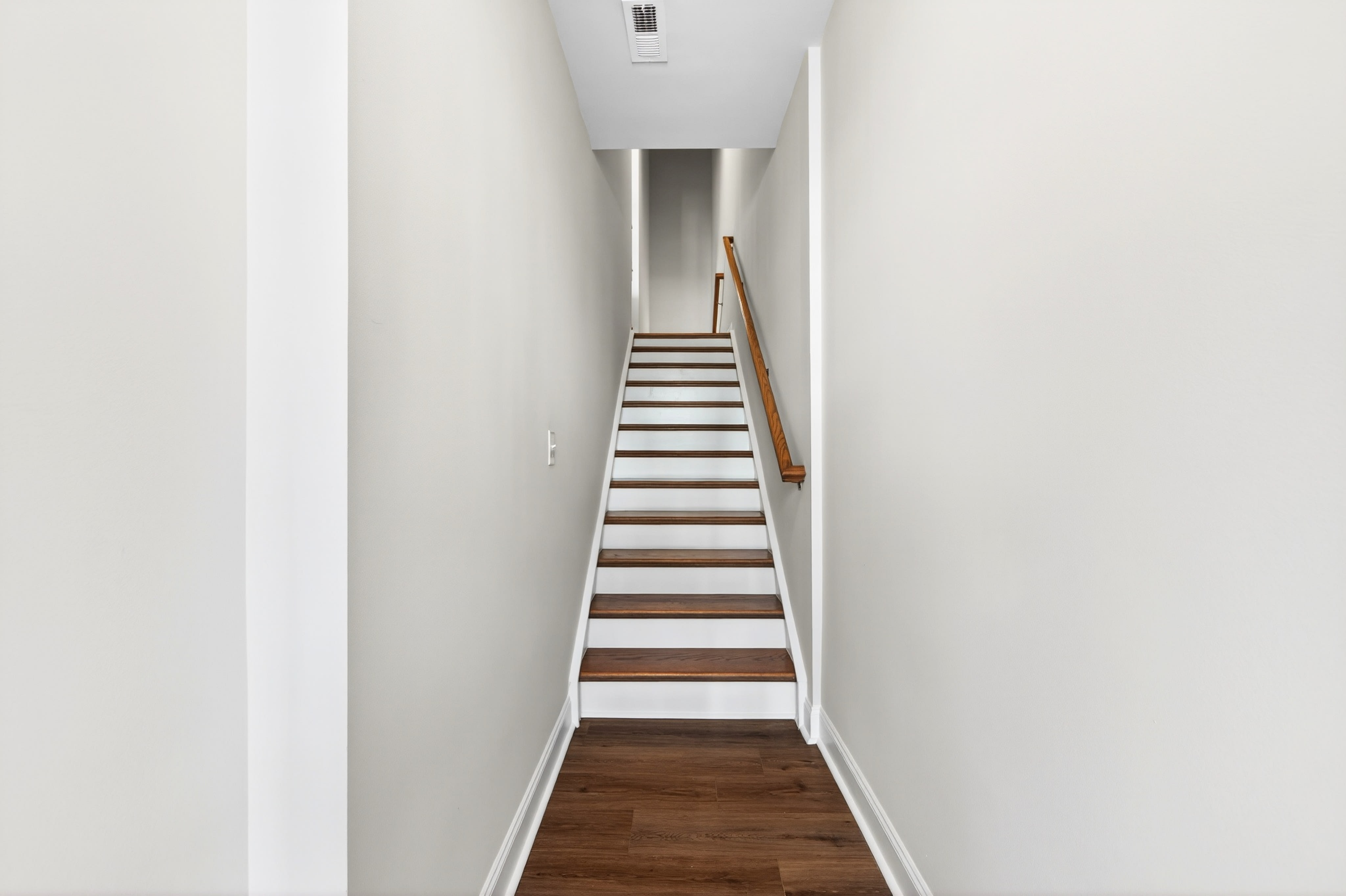 A long, narrow hallway with a wooden staircase leading upwards, surrounded by white walls and a light-colored floor.