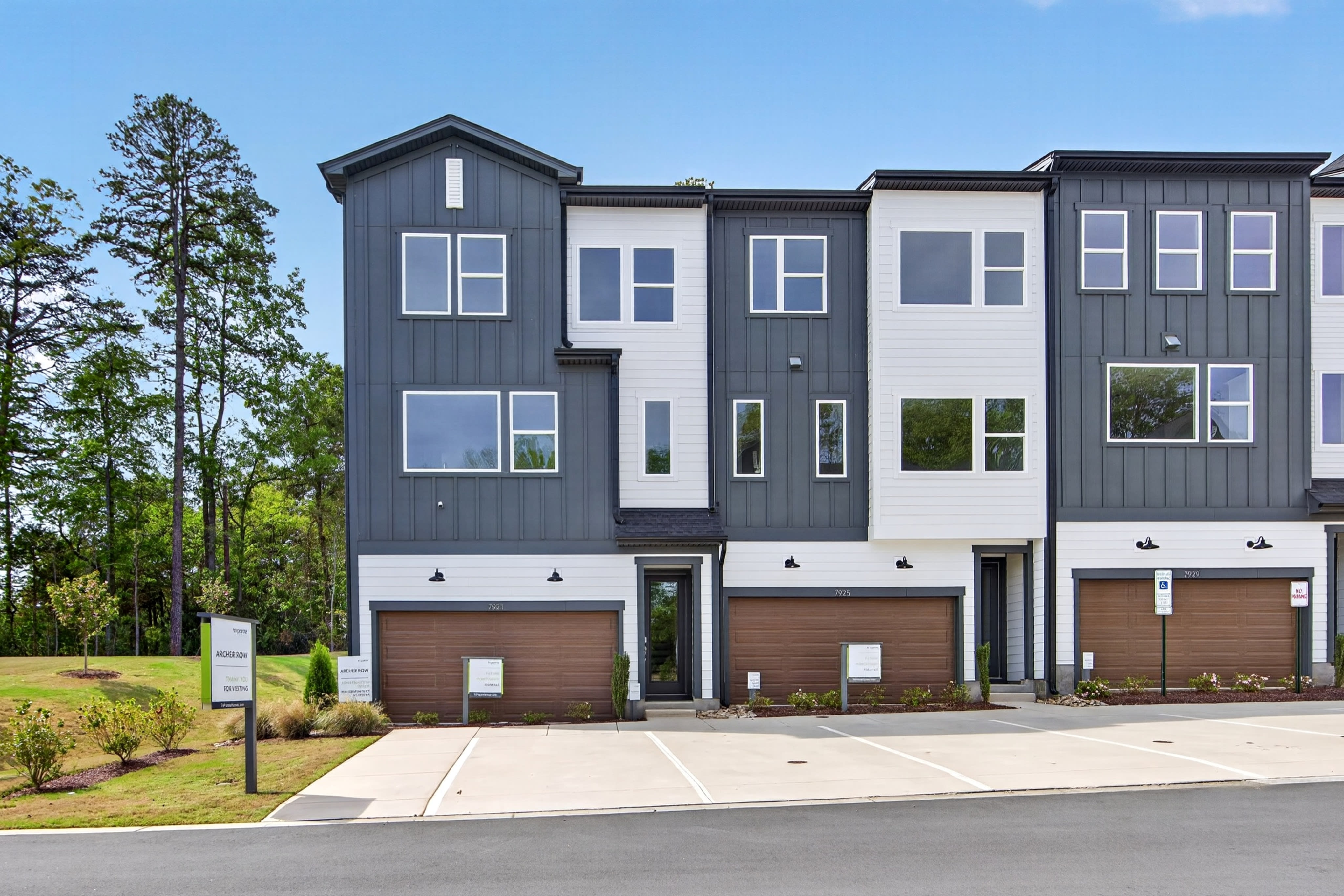 The image depicts a modern, multi-story townhouse complex with a gray and white exterior, surrounded by trees and a grassy area in the foreground.