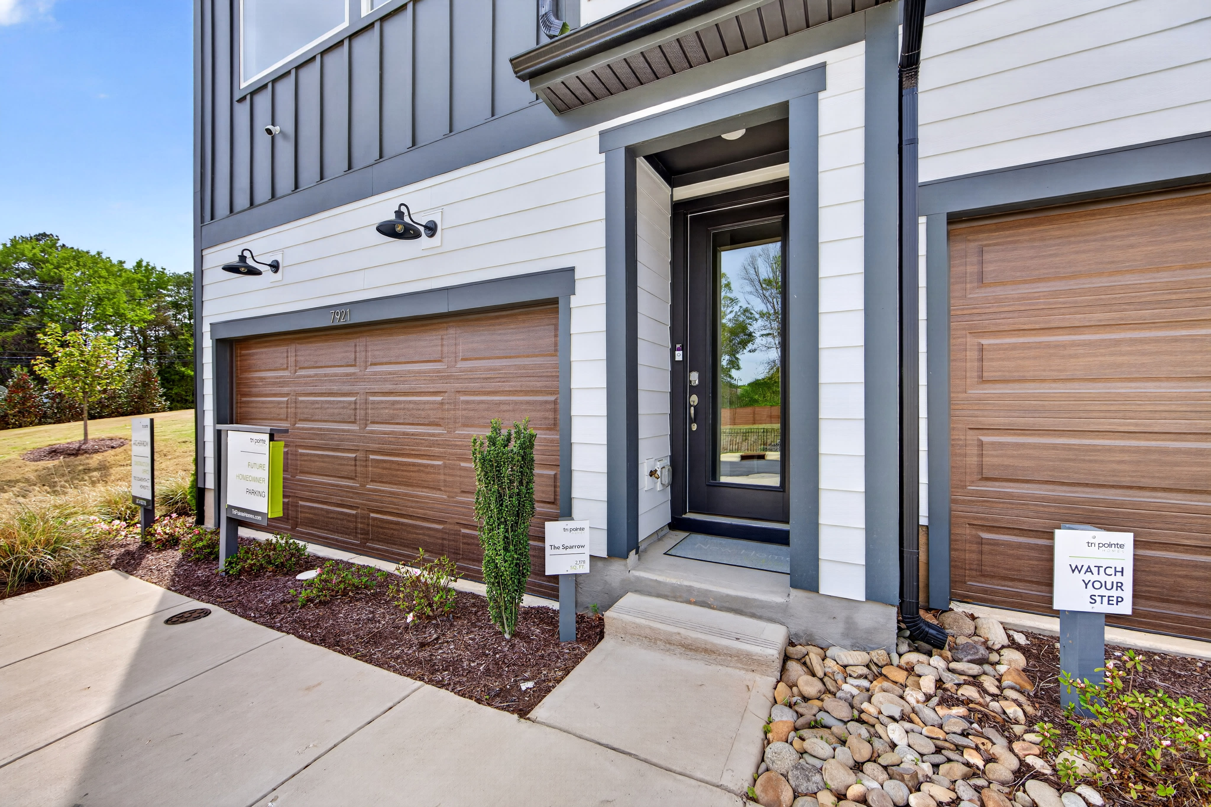 The image shows a modern, two-story building with a wooden garage door, a glass front door, and a landscaped pathway leading to the entrance. The building is surrounded by greenery and has a clean, contemporary design.