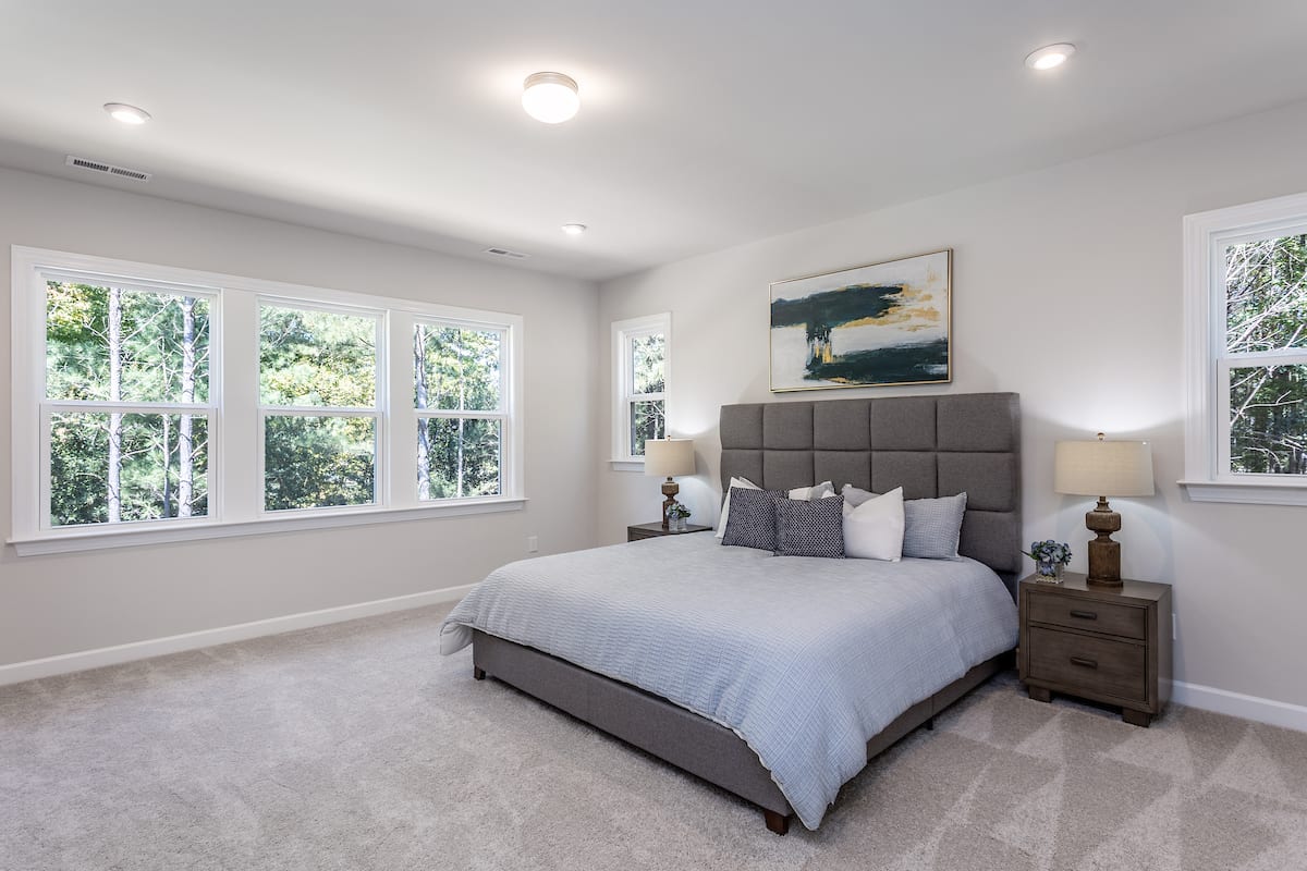 A spacious and well-lit bedroom with a large bed, a headboard, and a painting on the wall, surrounded by large windows overlooking a lush, green outdoor environment.