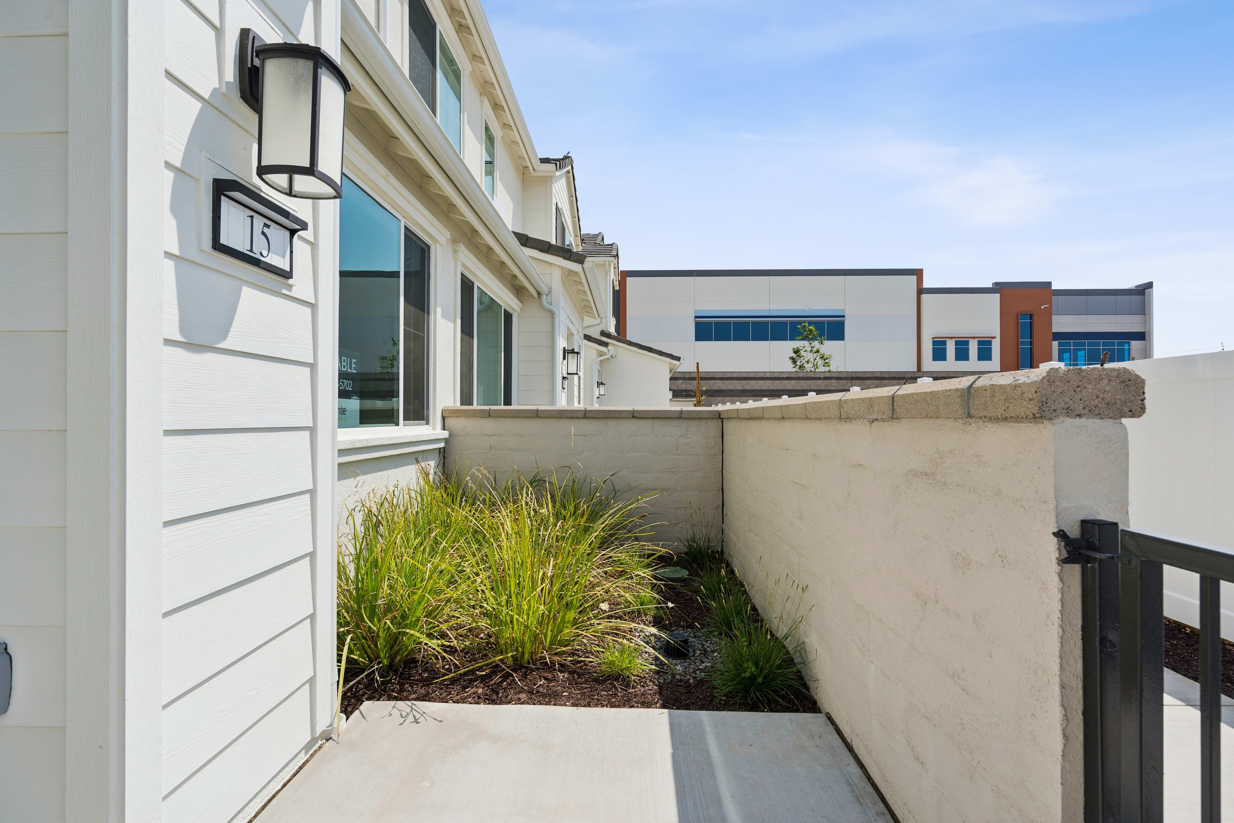 A modern building with a small garden area in the foreground, featuring tall grass and a concrete wall, set against a backdrop of additional buildings.
