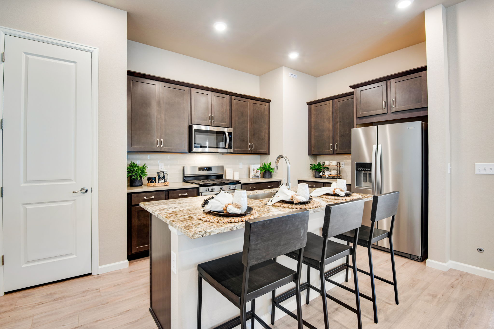A modern and well-equipped kitchen with dark wood cabinets, stainless steel appliances, and a granite countertop island with bar stools, set against a bright and airy backdrop.
