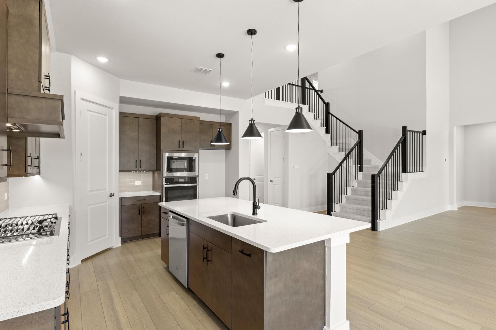 A modern, open-concept kitchen with a large island, stainless steel appliances, and a wooden staircase leading to the upper level in the background.