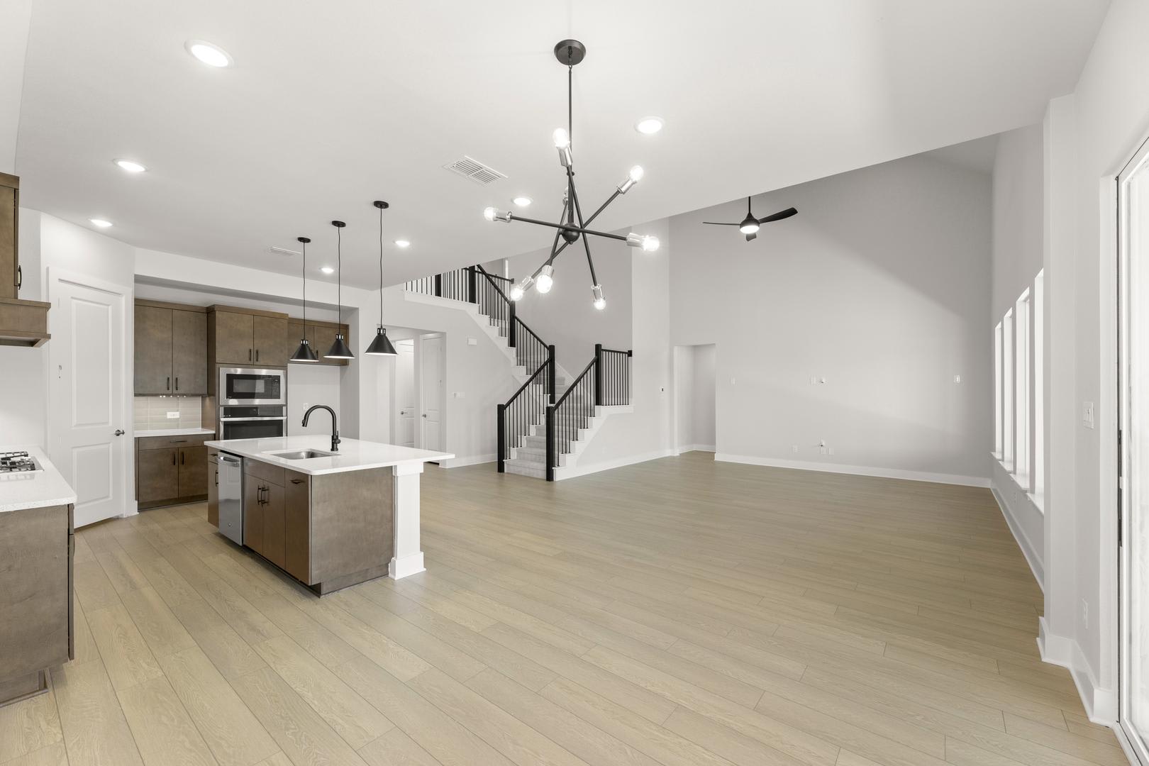 A modern, open-concept kitchen and living space with a sleek, minimalist design featuring light-colored hardwood floors, white walls, and a striking black chandelier overhead.
