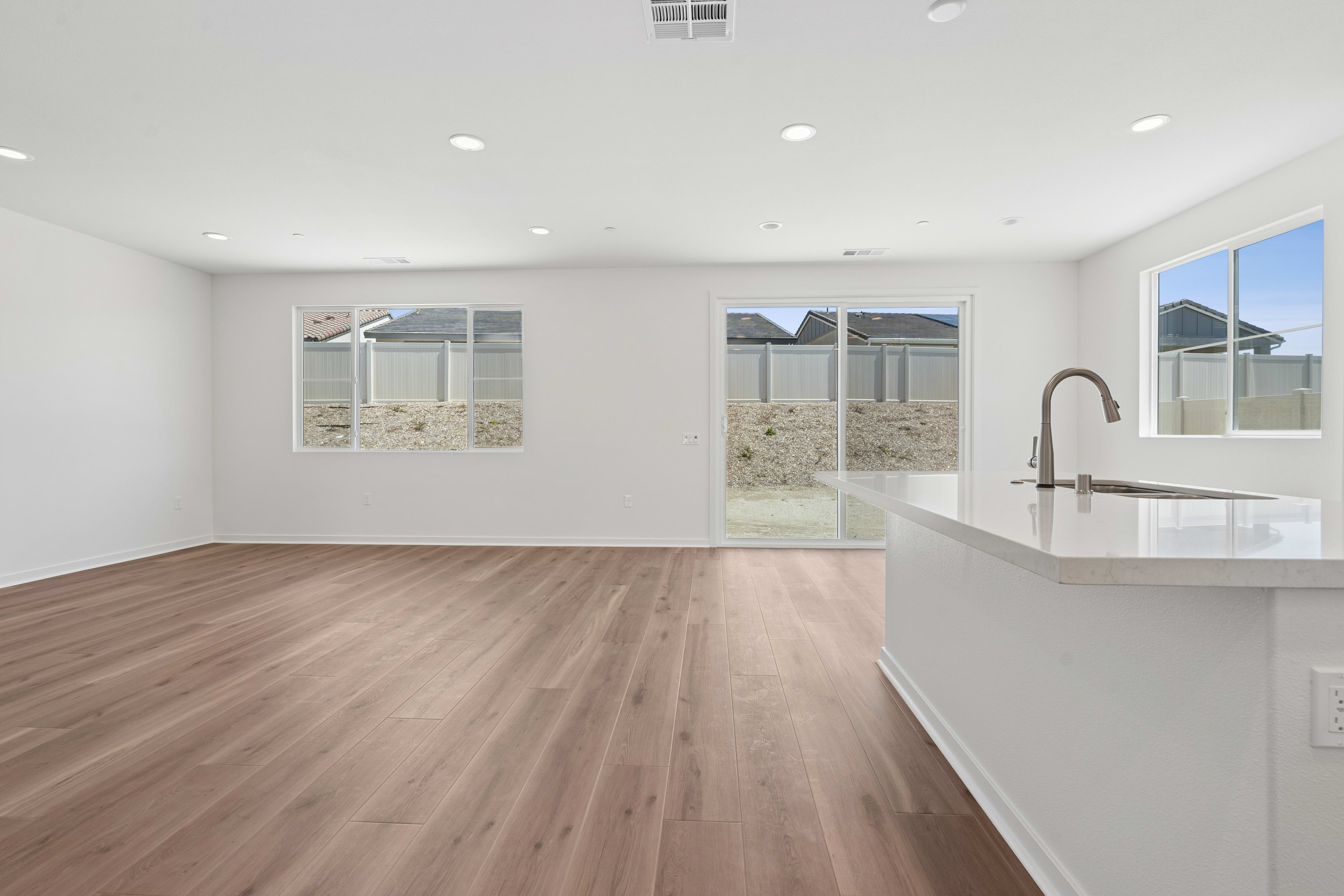 A spacious, open-concept living area with hardwood floors, white walls, and large windows providing natural light. The kitchen counter and sink are visible in the foreground.