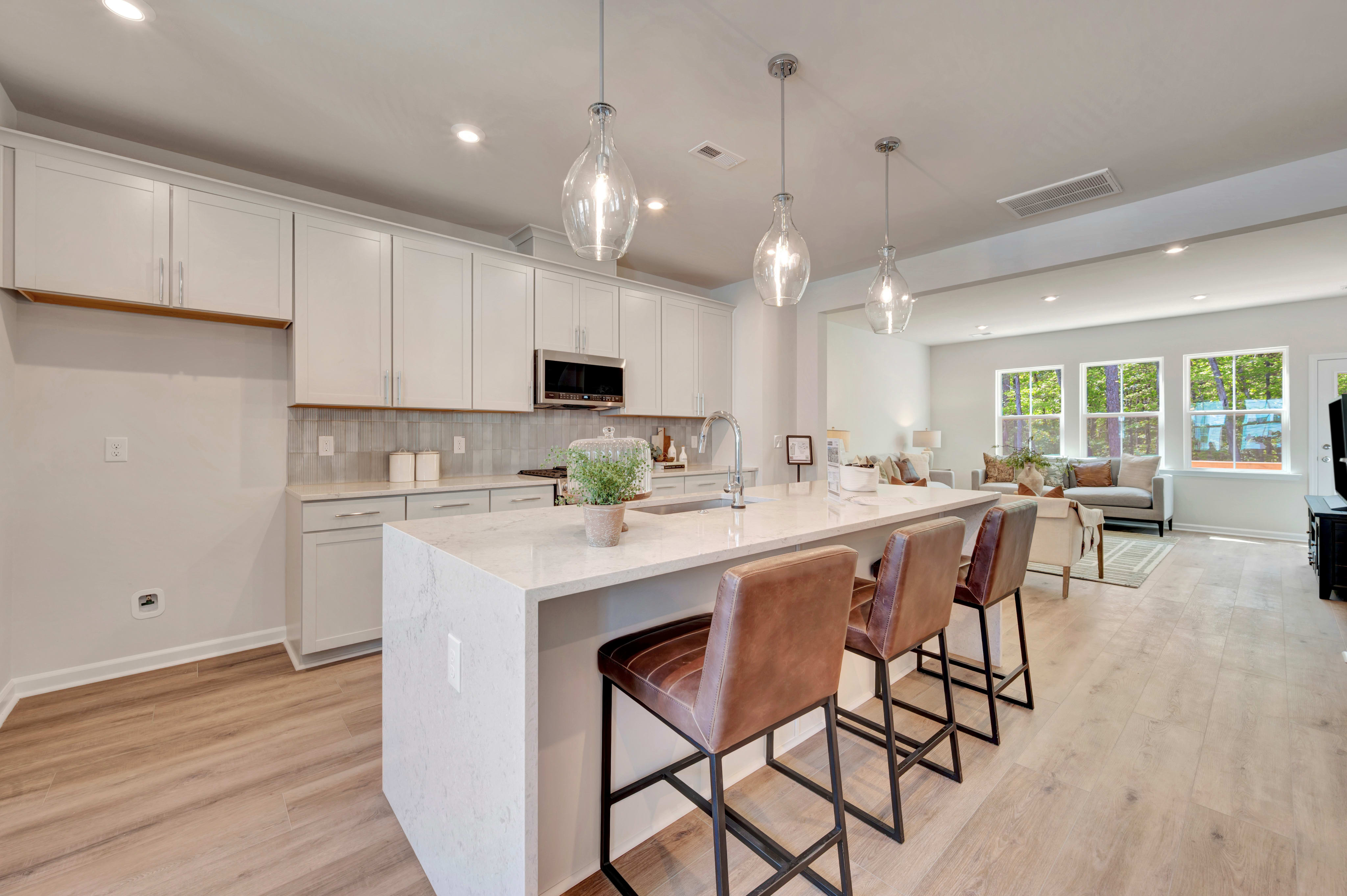 A modern and bright kitchen with white cabinets, a large island with bar stools, and pendant lights hanging from the ceiling, surrounded by large windows that let in natural light.