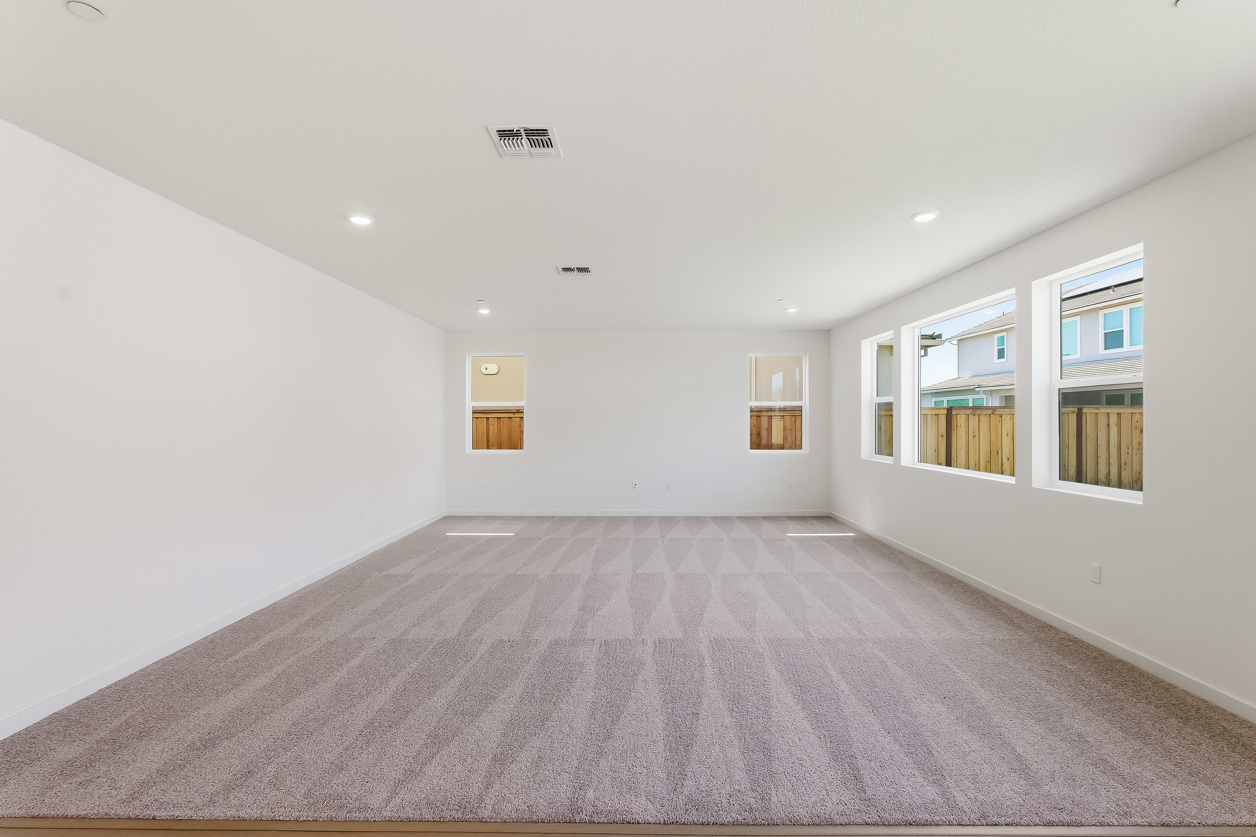 An empty, spacious room with white walls, a gray carpeted floor, and two windows providing natural light.