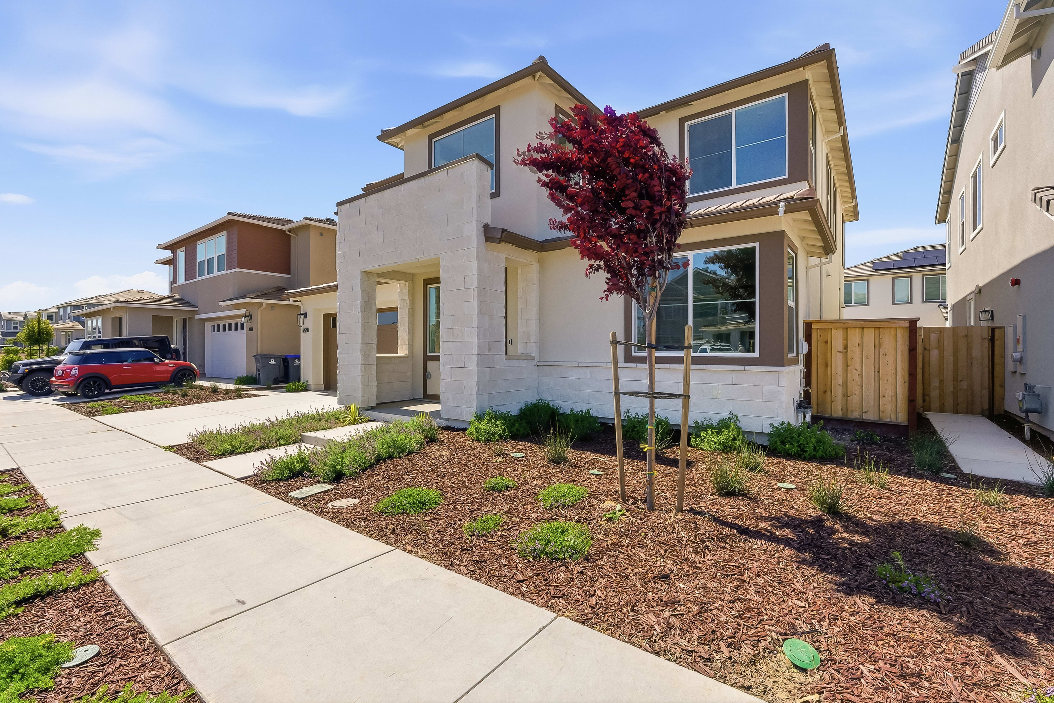 A modern two-story residential building with a well-landscaped front yard, featuring a blooming tree and a neatly maintained garden, set against a clear blue sky.