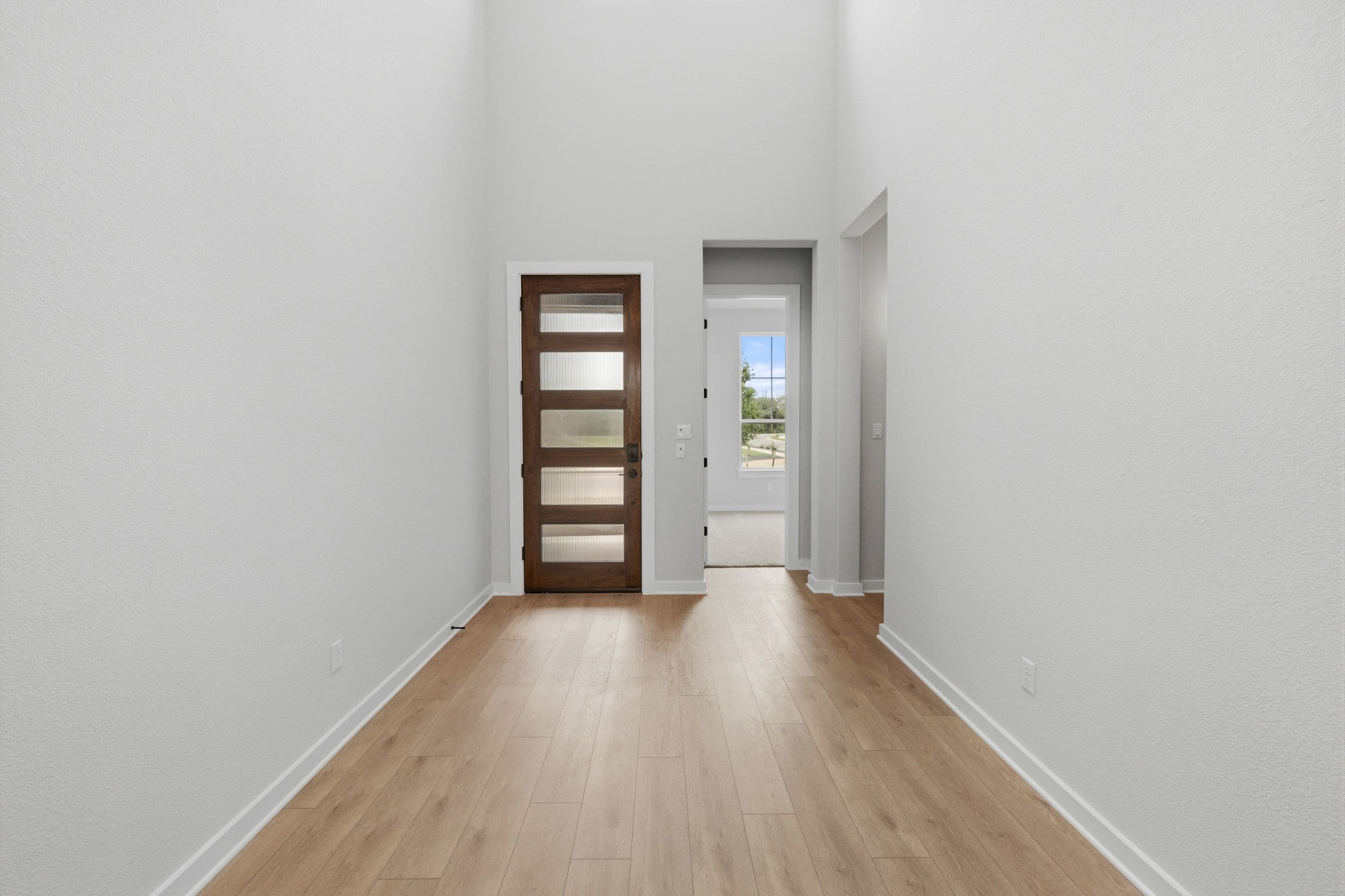 A long, narrow hallway with white walls, a wooden door, and a wooden floor leading to an open doorway with a view of the outdoors.