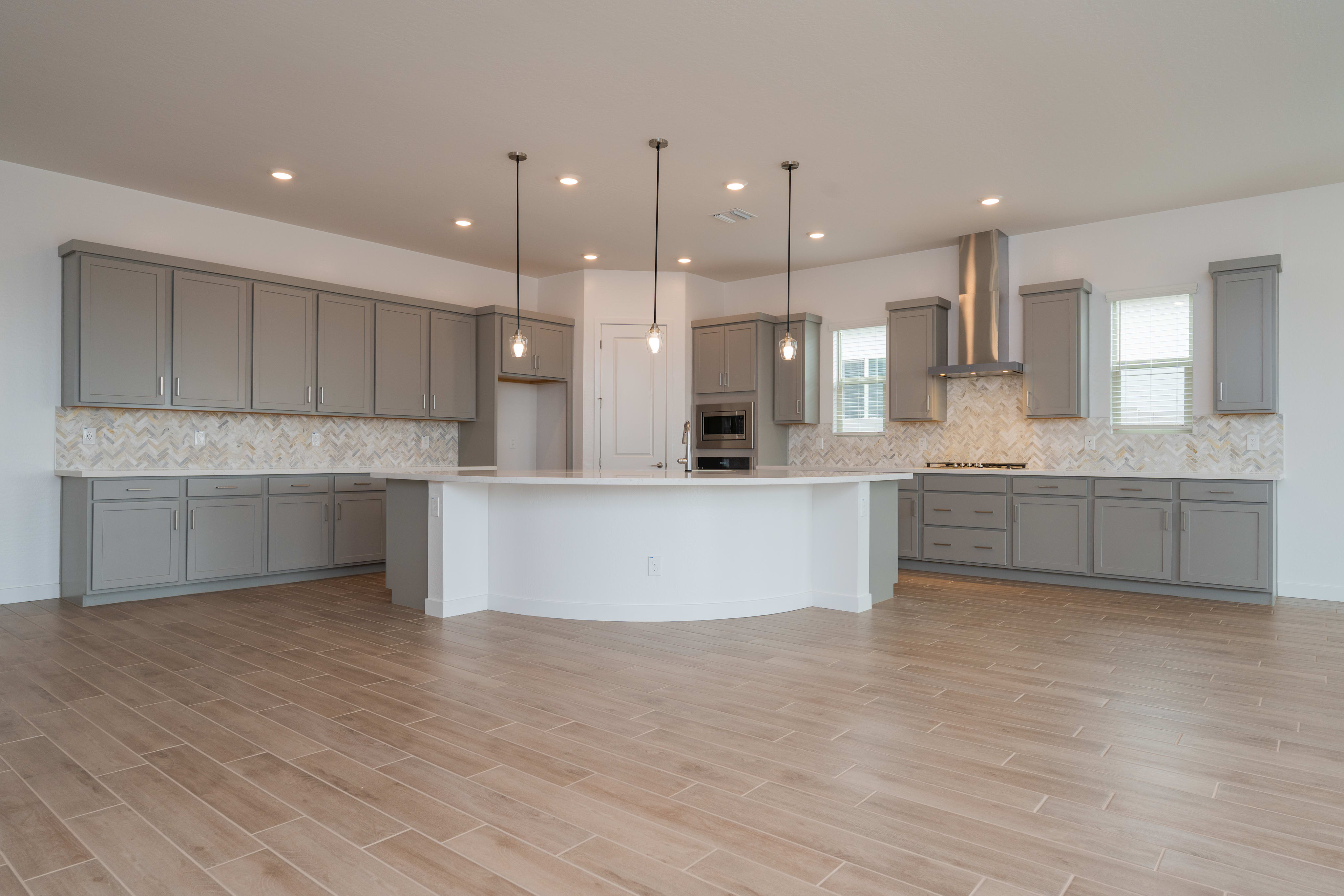 A spacious and modern kitchen with gray cabinets, a white countertop, and a hardwood floor. The room is well-lit with recessed lighting and pendant lamps.