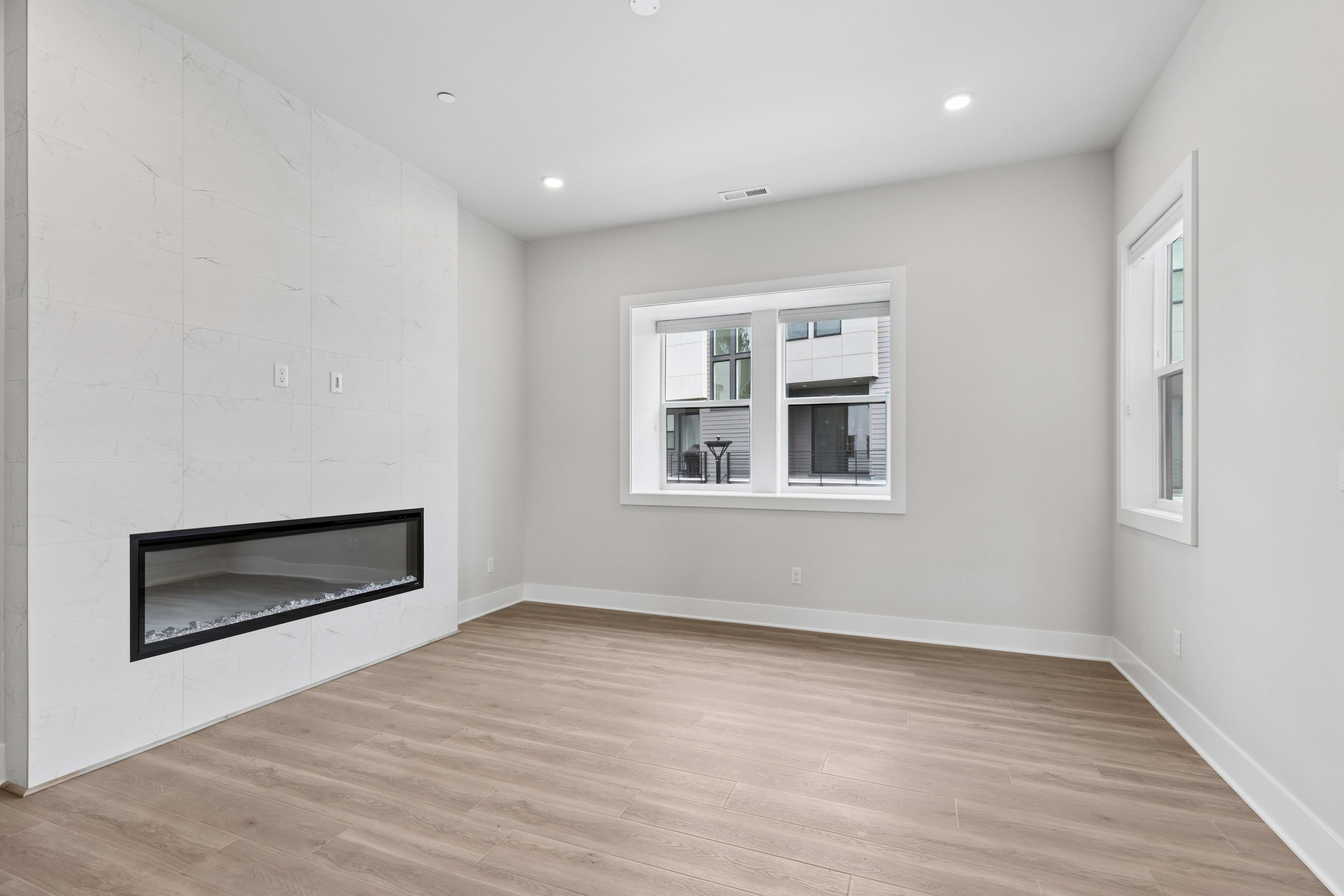 A minimalist, well-lit room with a fireplace and hardwood flooring, featuring a large window that provides natural light.