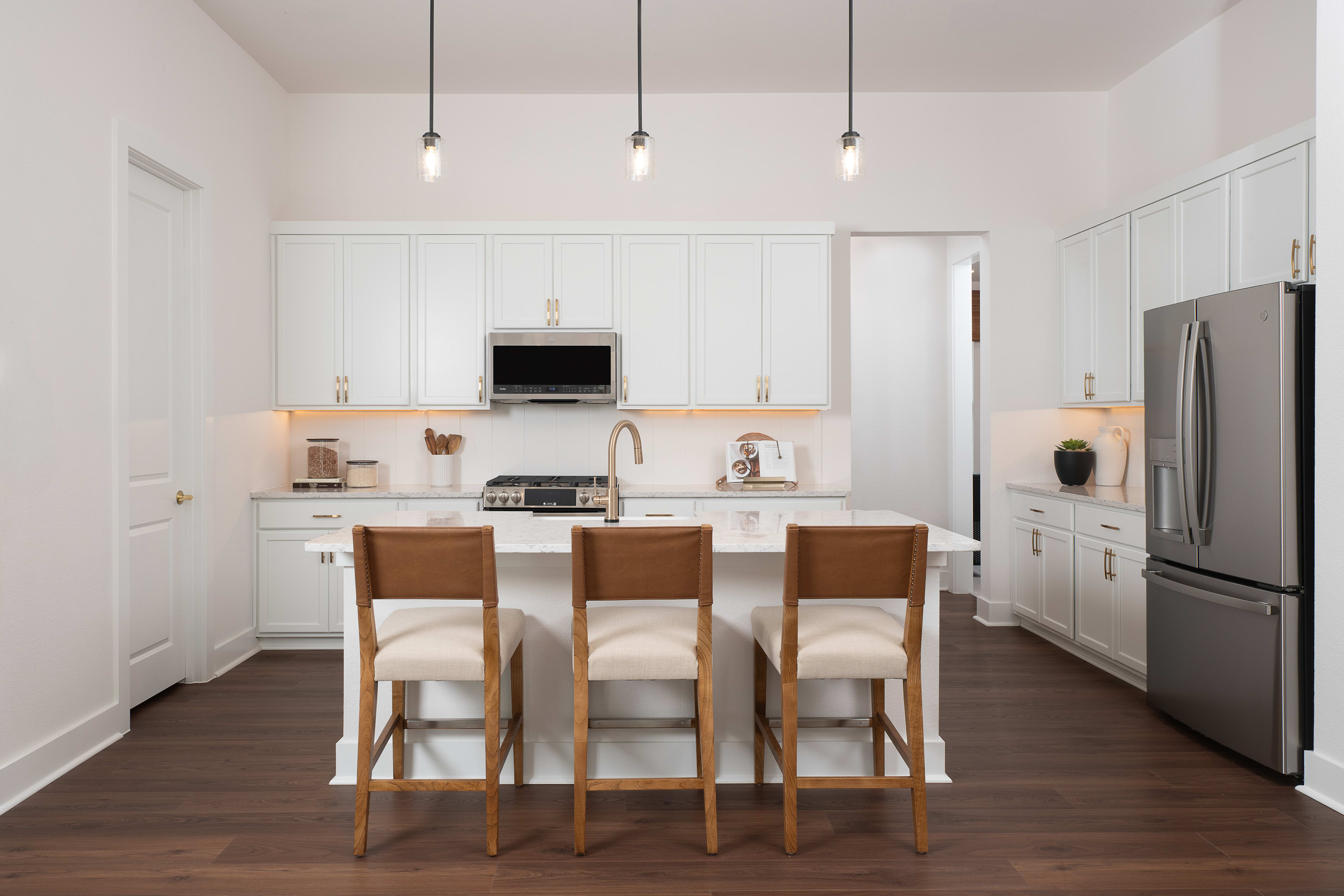A modern, minimalist kitchen with white cabinets, wooden bar stools, and pendant lighting, set against a hardwood floor.