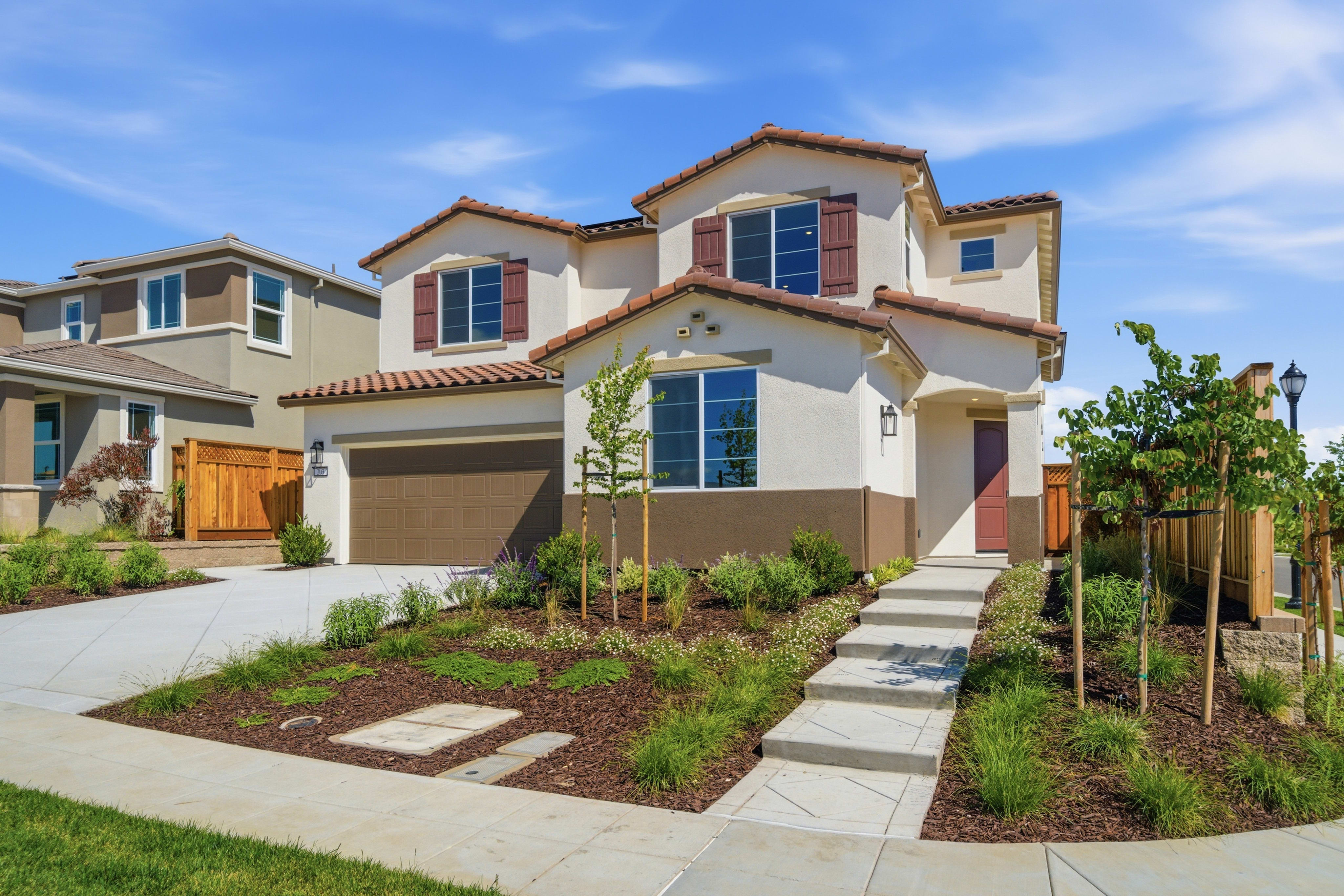 A two-story Mediterranean-style house with a tiled roof, surrounded by a well-landscaped yard featuring a paved walkway, lush greenery, and a clear blue sky in the background.