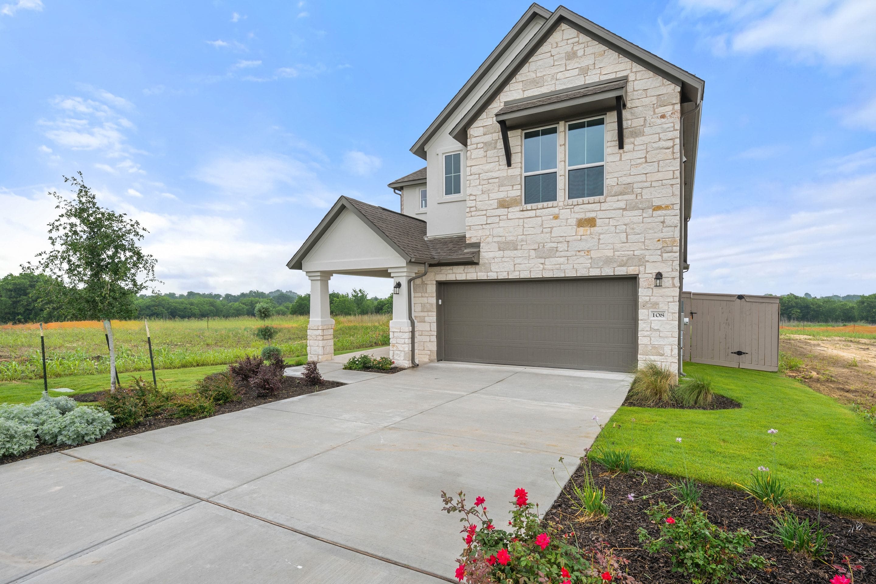 A modern two-story house with a stone exterior, a garage, and a well-maintained landscaped yard with flowers in the foreground, set against a backdrop of a grassy field and a blue sky with clouds.