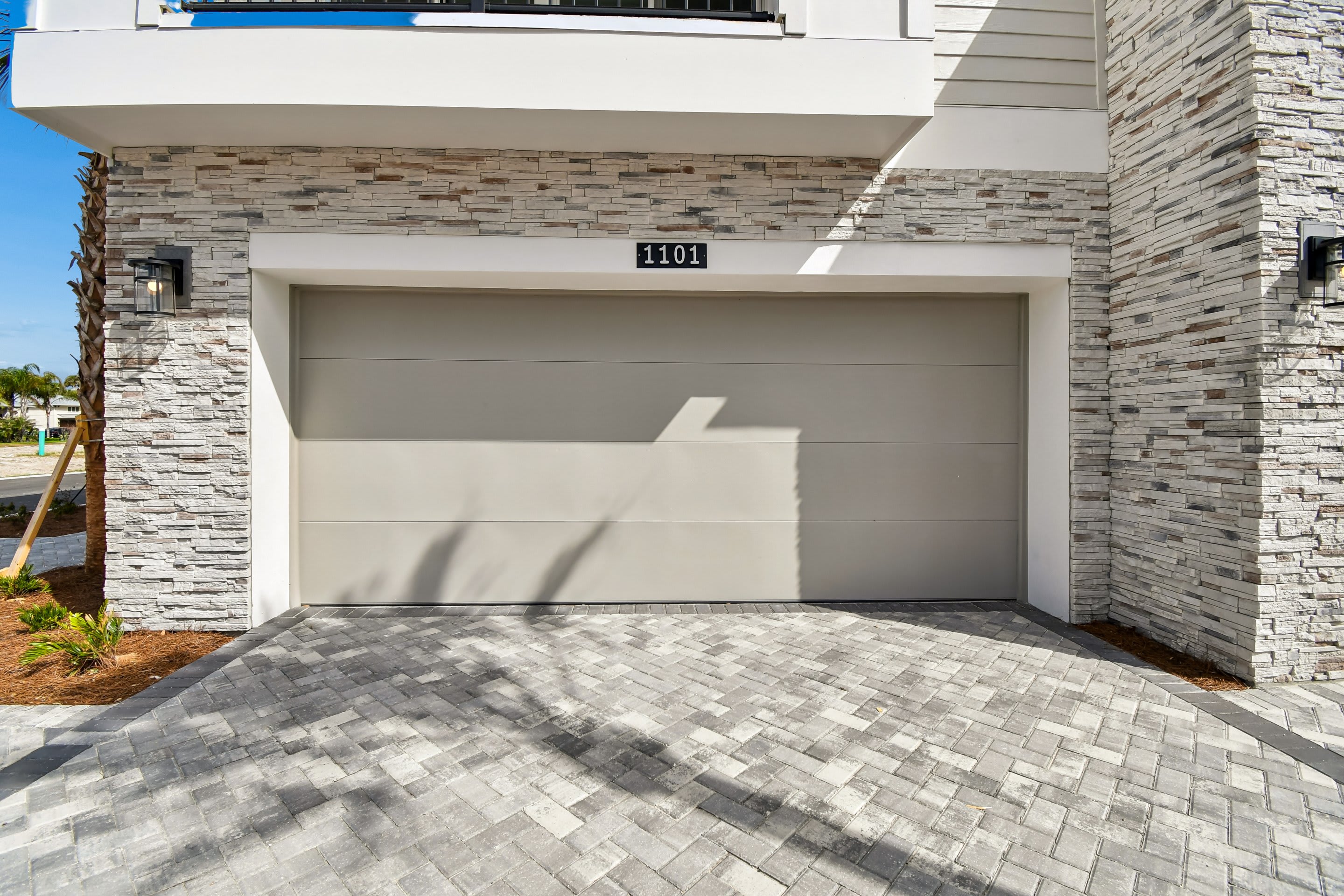 A modern garage door with a geometric design is centered in the frame, set against a stone-clad facade, with a patterned paver driveway in the foreground and a clear blue sky in the background.