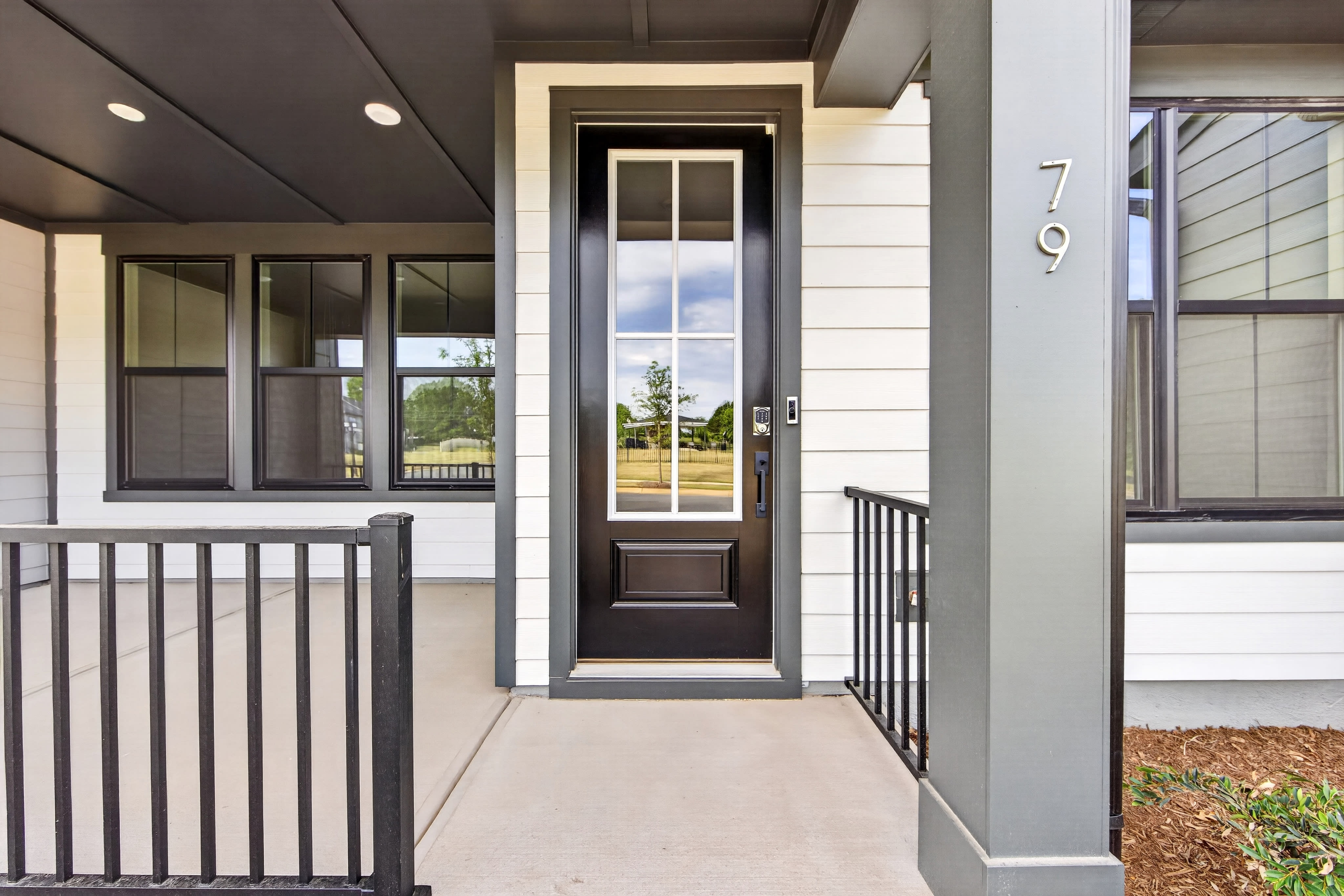 A modern front porch with a black door and windows is visible in the foreground, while a white-sided house and a glimpse of greenery form the background.