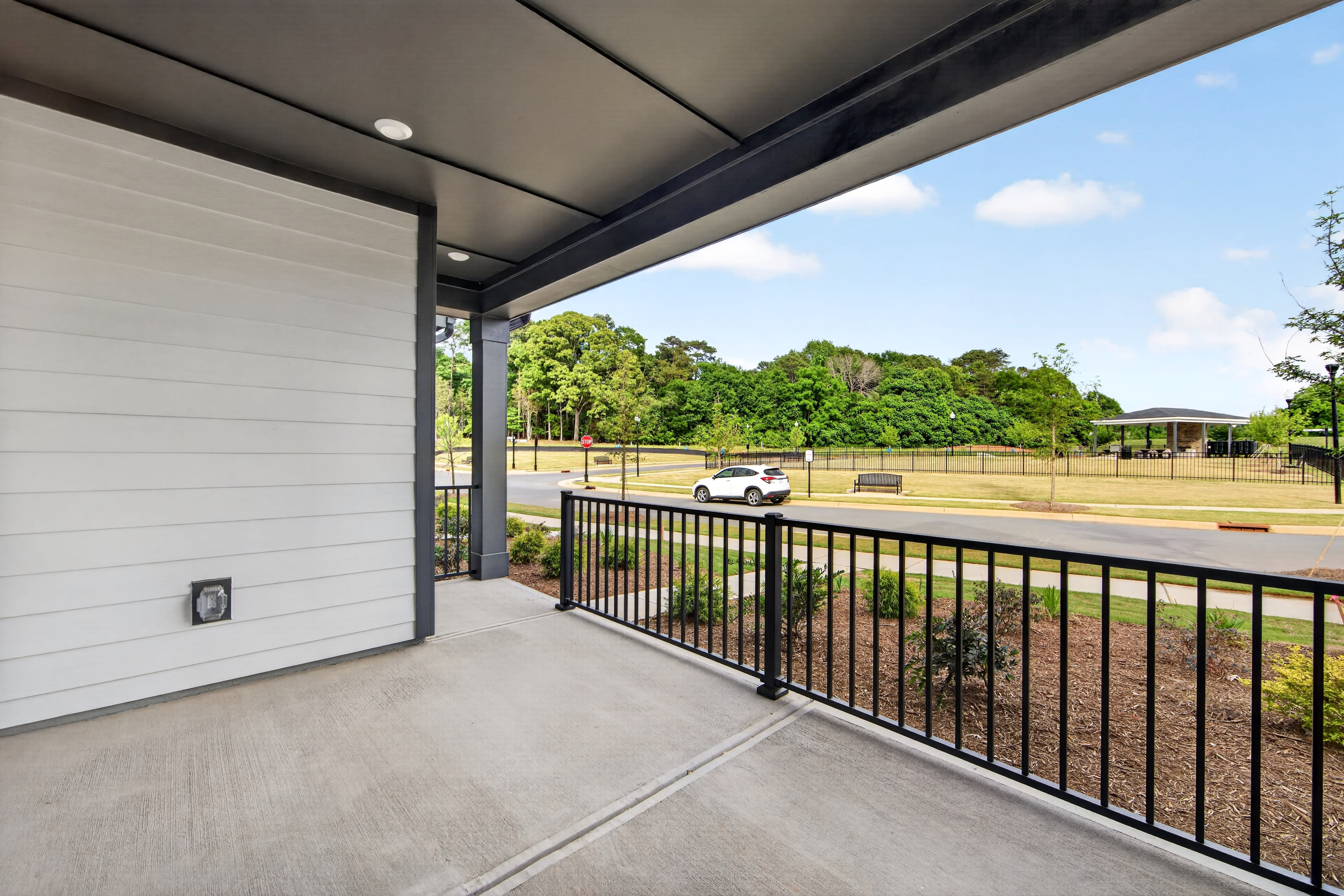 A modern porch with a white wall and black railing overlooks a grassy park with trees and a white car.