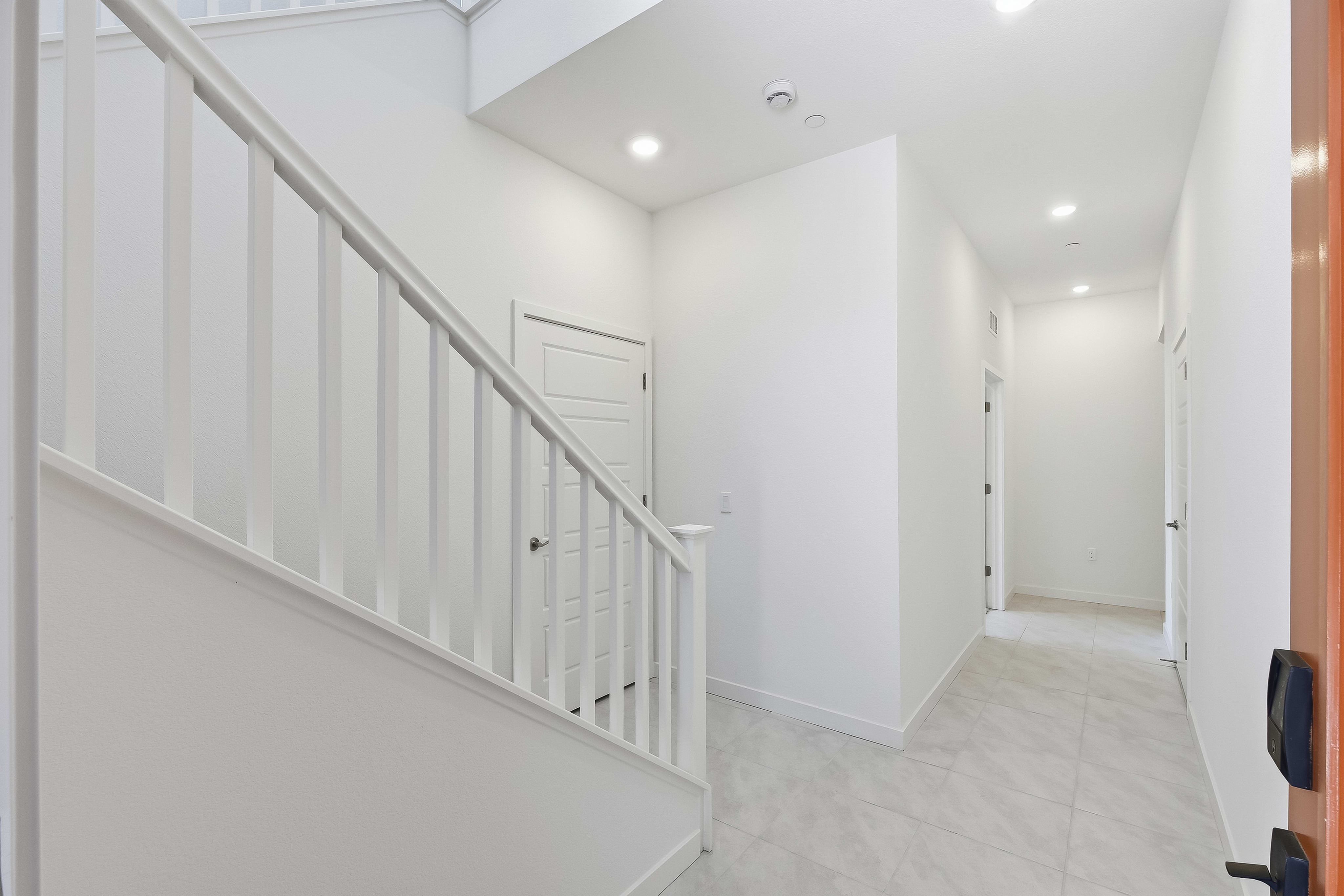 A white staircase with a railing dominates the foreground, leading into a bright, modern hallway with tiled floors and several doors in the background.