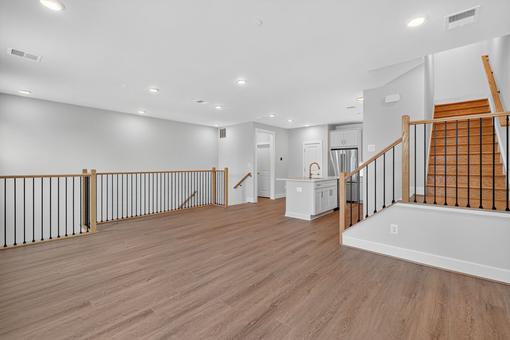 A spacious, modern living area with light wood floors in the foreground leads to a kitchen with a white island and stainless steel appliances in the background, all under a bright, well-lit ceiling.