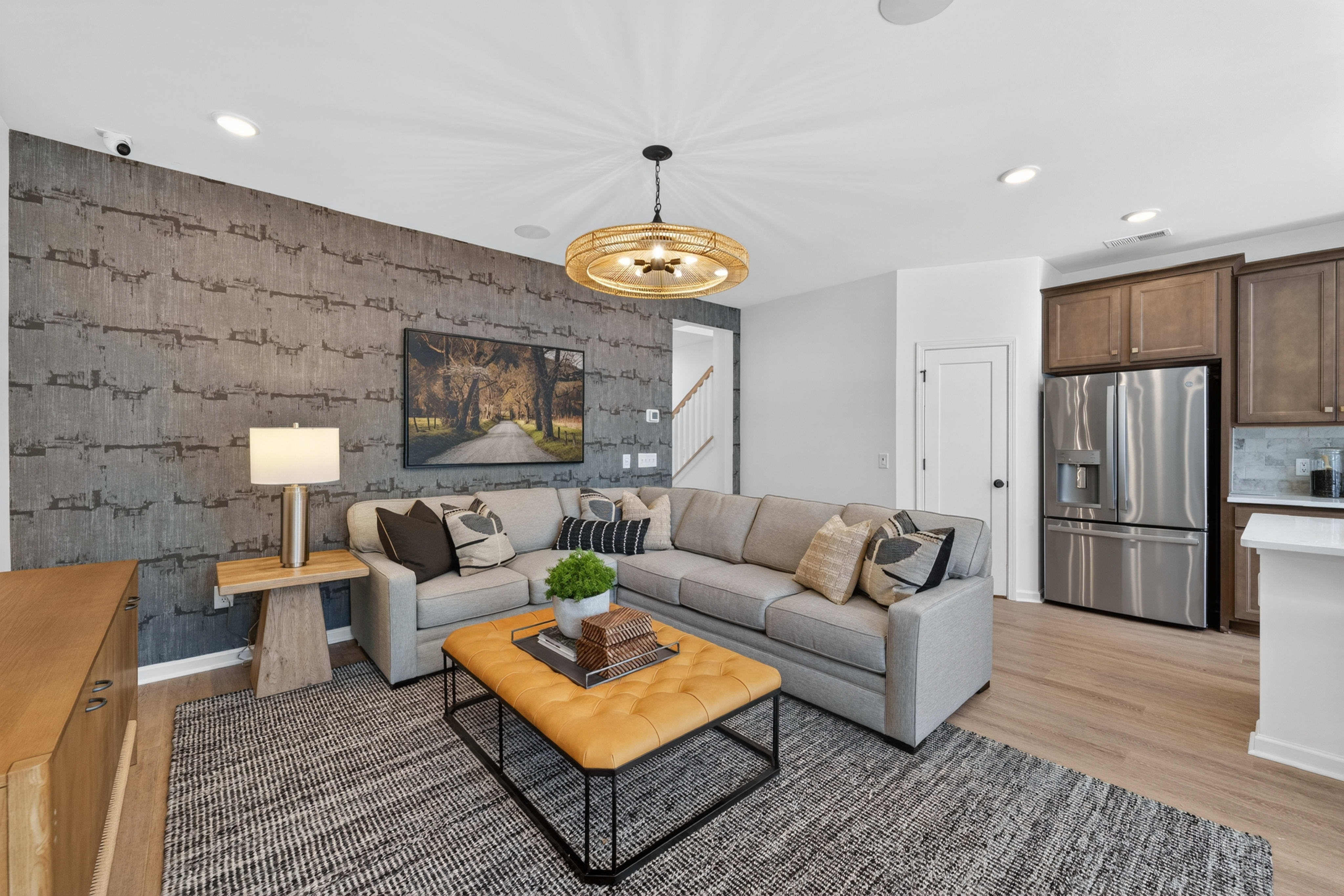 A modern living room with a gray sectional sofa and a tufted ottoman in the foreground, and a stainless steel refrigerator and kitchen cabinets in the background.