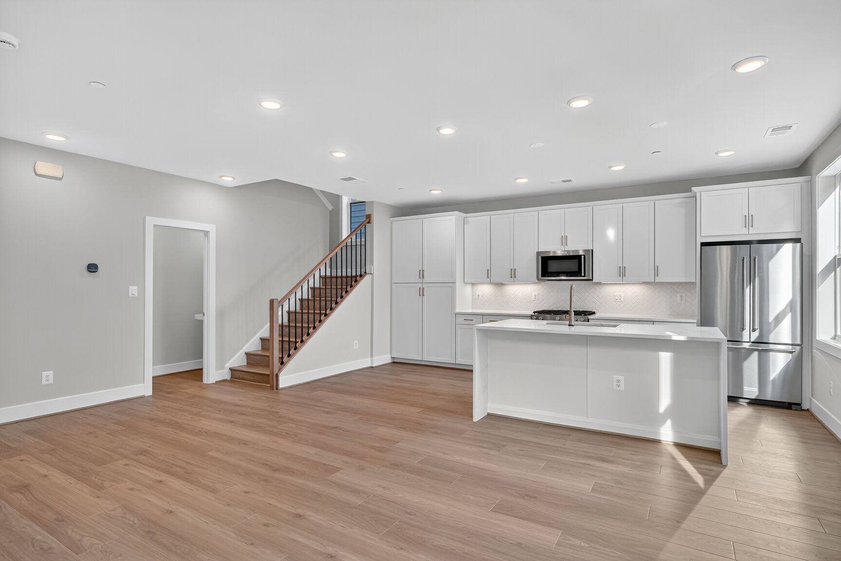 A modern, open-concept living space features a light wood floor in the foreground, leading to a sleek white kitchen with stainless steel appliances and a staircase in the background.