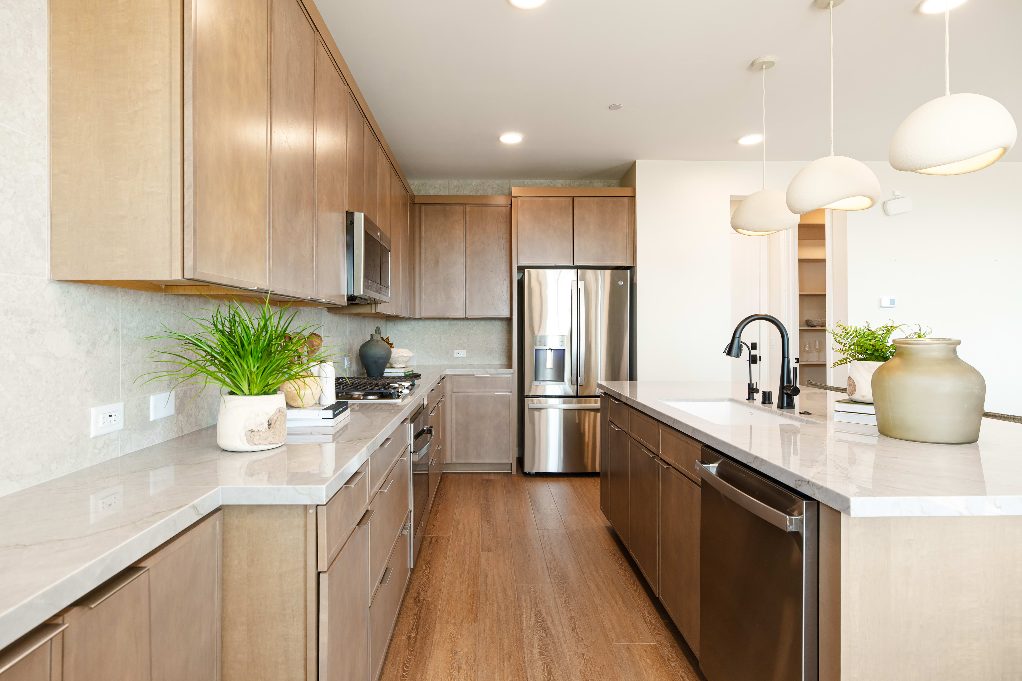 A modern kitchen with light wood cabinets and stainless steel appliances features a white countertop with plants in the foreground and a bright, open space in the background.