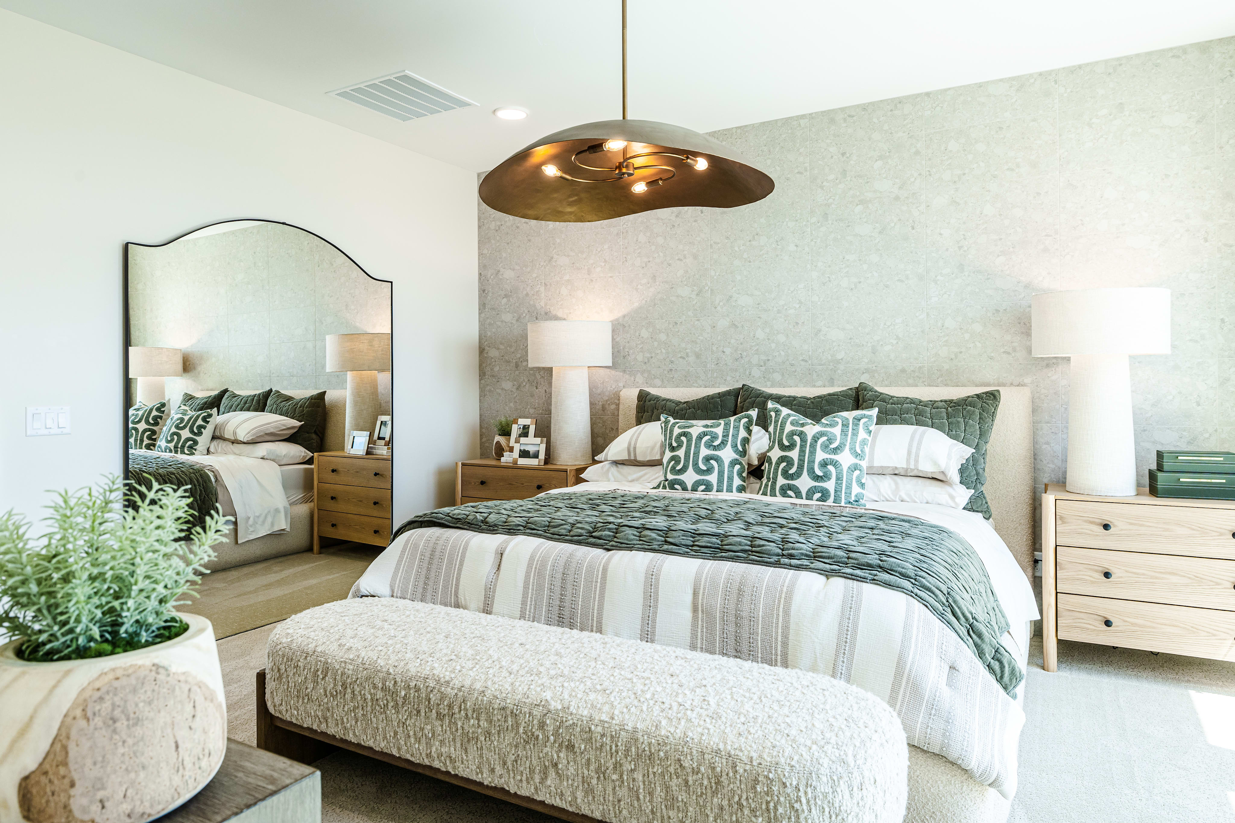A plush bench sits in front of a king-sized bed with patterned pillows, while a large mirror reflects the room and a textured wallpaper adorns the wall behind the bed.