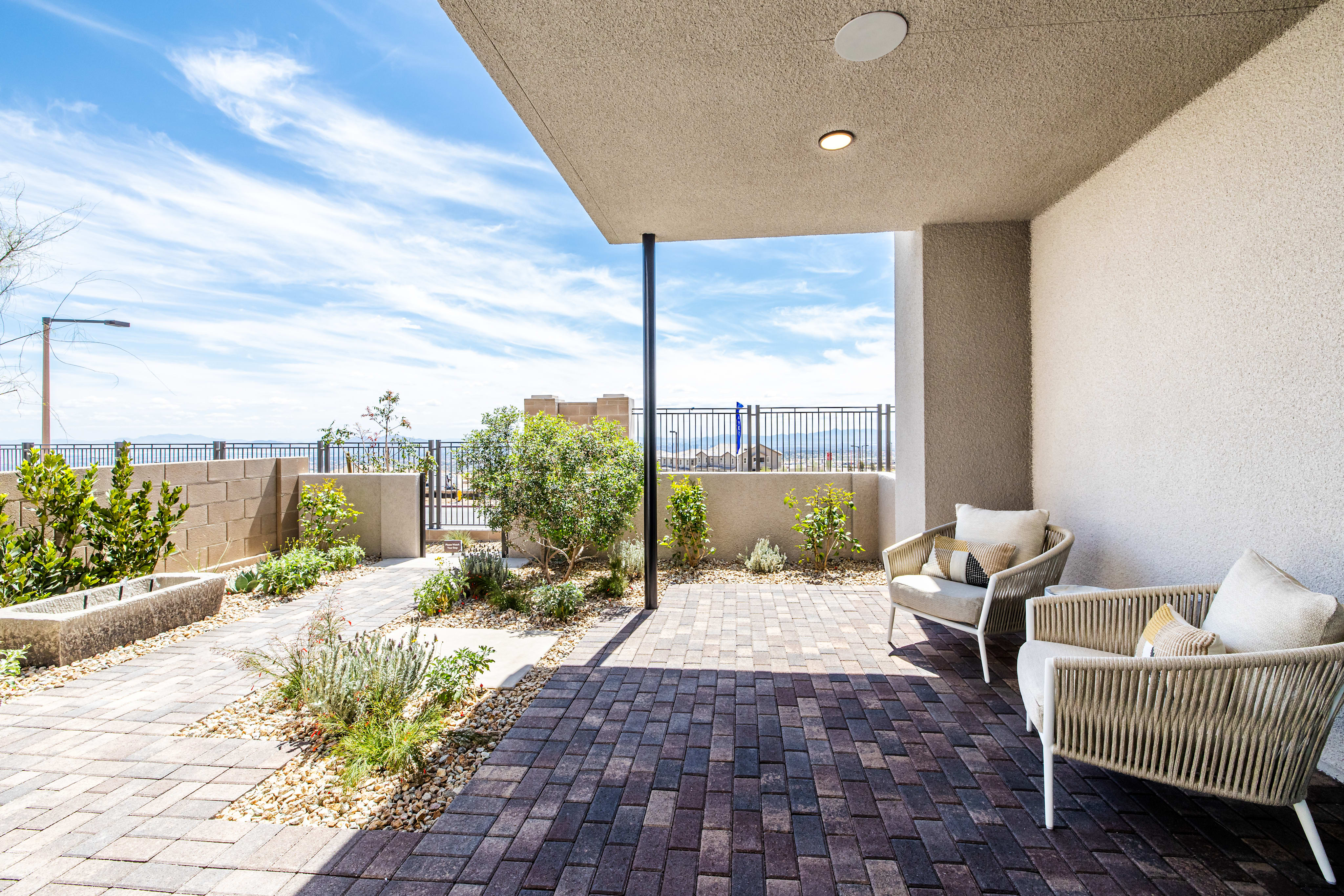 Two chairs sit on a paved patio in the foreground, with a landscaped yard and distant hills under a bright blue sky in the background.